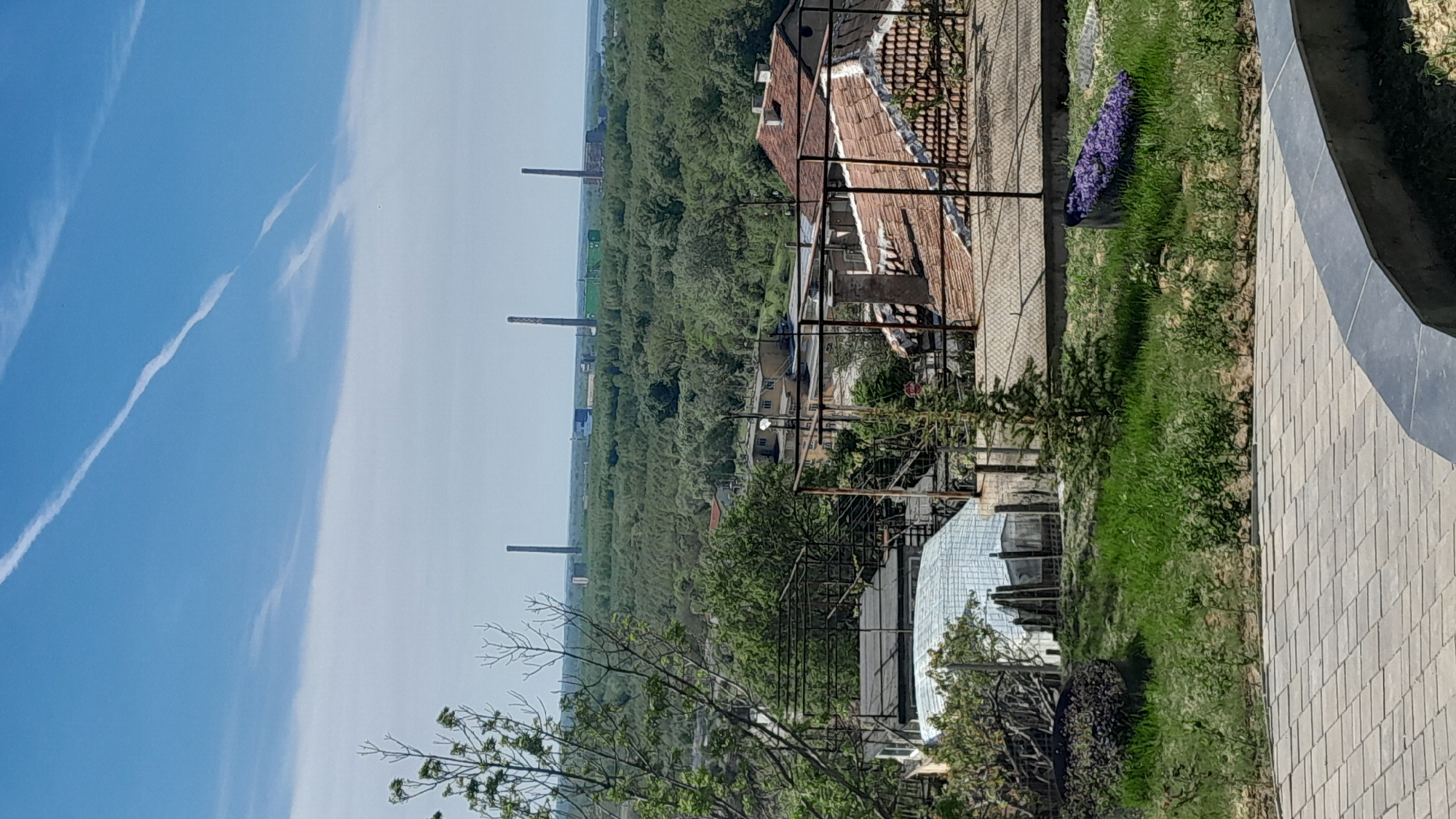 The image depicts a view from a raised position overlooking the city of Svishtov, Bulgaria. In the distance, industrial smokestacks are visible among a dense expanse of trees. Closer to the viewer, there are older buildings with red tile roofs and metal railings. The foreground features a paved walkway and a grassy area with landscaping, including purple flowers. The sky is blue with contrails.