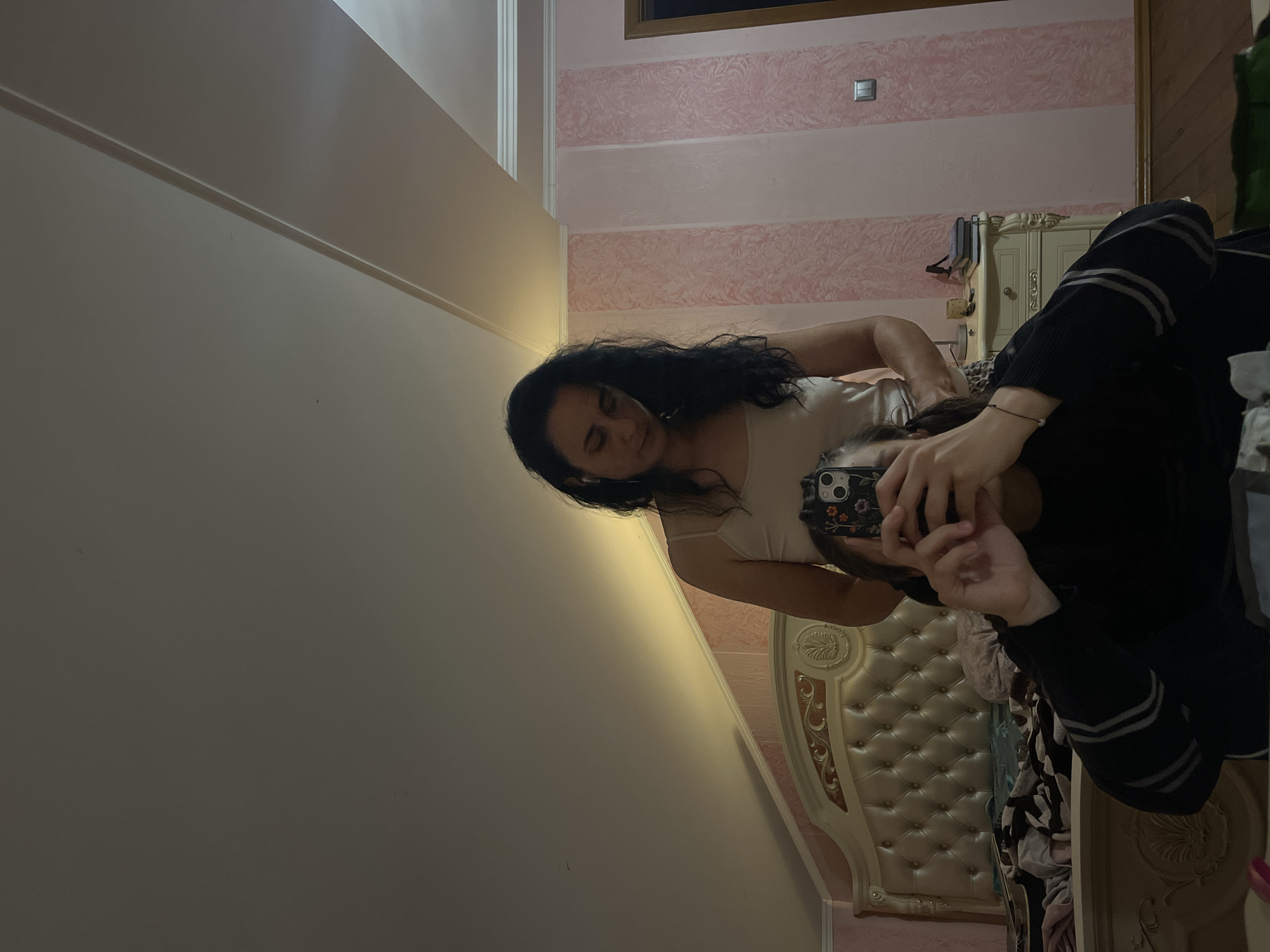 An indoor, dimly lit scene in a room identified as being in Tarnivtsi, Ukraine, captures two individuals via a mirror reflection. The image depicts an adult female with long, dark, wavy hair, wearing a light-colored tank top and a white earbud in her left ear, standing and looking downwards. Her lower body is partially obscured by another individual and patterned fabric. In the foreground, a second person is seated or crouched, holding a silver smartphone with a floral-patterned case to take the selfie. This individual is wearing a dark long-sleeved top with light stripes on the sleeves and a thin dark bracelet with a white bead on the left wrist.
The room features a prominent, ornate cream-colored bed with a large tufted headboard and decorative wooden accents. Disheveled bedding, including black and white patterns, is visible on the bed. To the right of the bed is a cream-colored nightstand with carvings, upon which a small lamp, a brown cup, and stacked notebooks are present. The right wall is adorned with alternating vertical stripes of two textured pink wallpapers and a white crown molding. A white double light switch is mounted on this wall. The ceiling is sloped, light-colored, and includes a visible structural beam. Warm light emanates from the left, casting a yellowish glow on the ceiling and upper walls. A dark wooden floor and a partial dark green mat are visible in the lower right. A dark brown doorframe is seen in the far right background.