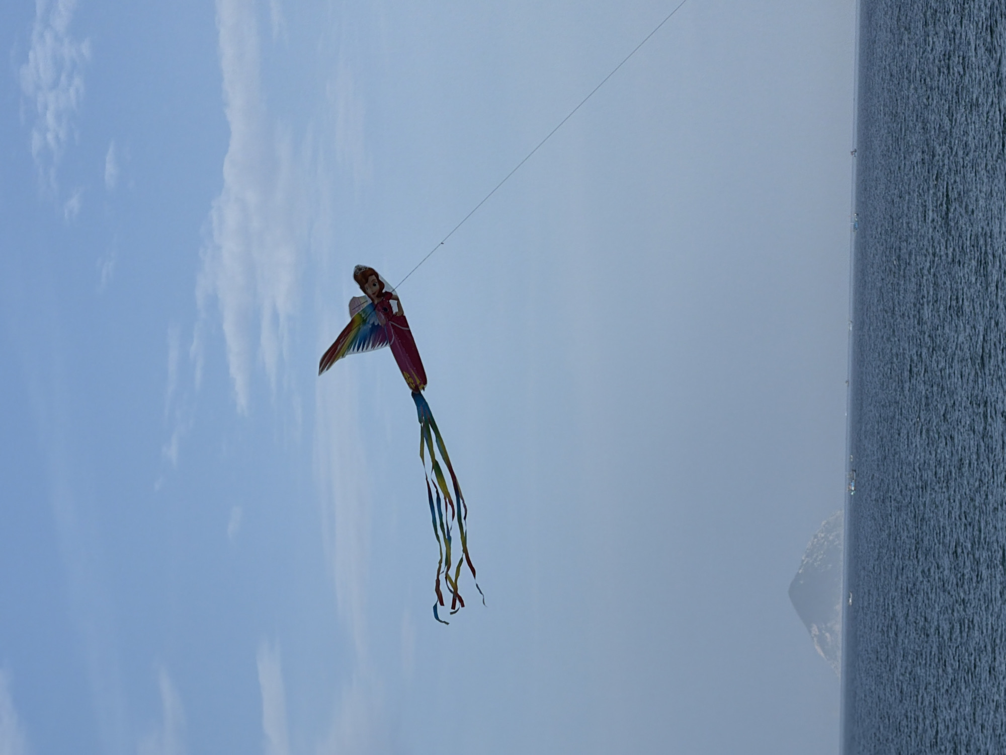 A colorful kite shaped like a winged doll flies high in a pale blue sky over the ocean. The kite has rainbow-colored wings and a pink dress. Multiple ribbon-like tails trail behind it. In the distance, a small, dark, conical island rises from the sea, and several small boats are scattered across the water. The image appears to have been taken from a coastal area in or around Nha Trang, Vietnam, given the presence of the island and the style of the boats.