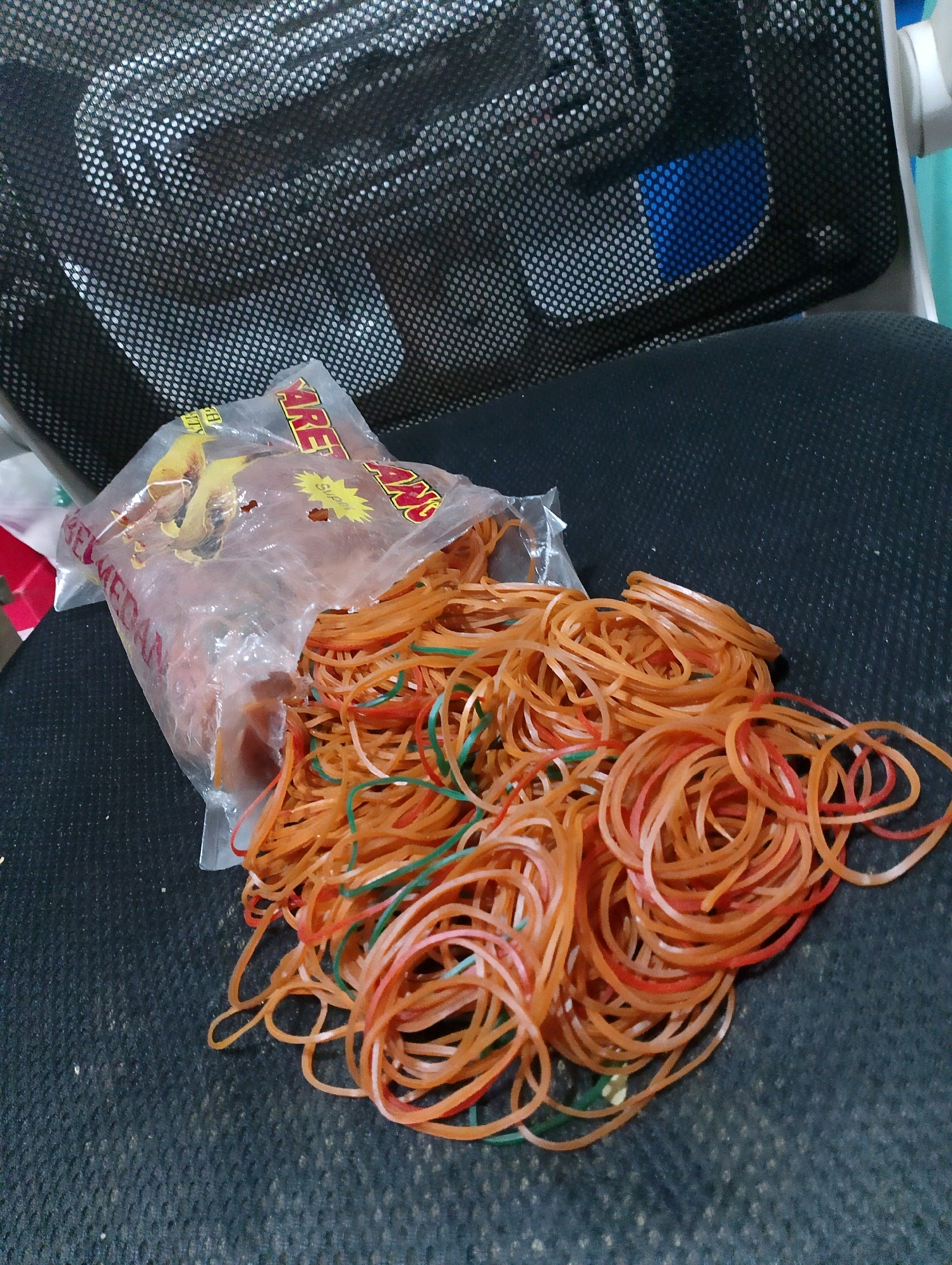 A spill of orange and green rubber bands lies on a textured black surface, likely a chair or fabric. A torn plastic bag, partially filled with more rubber bands, is to the left of the spill, with visible text on the bag. In the background, a black mesh basket with a blue object inside is visible. The scene is indoors. The location is not specified in the image.