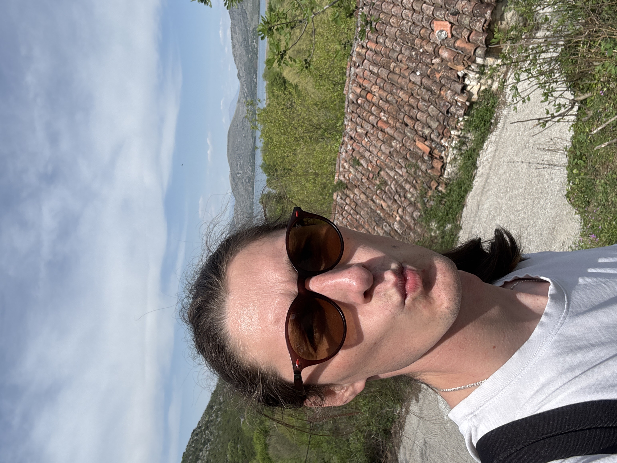 The photograph shows a selfie of a fair-skinned person with sunglasses, a white t-shirt, and dark hair, in an outdoor location. The person is facing the camera with a slightly puckered expression. The background includes a tiled rooftop, green vegetation, a visible mountain range, and a cloudy sky. Given the prompt, the location is identified as the city of Bistrice, Montenegro.