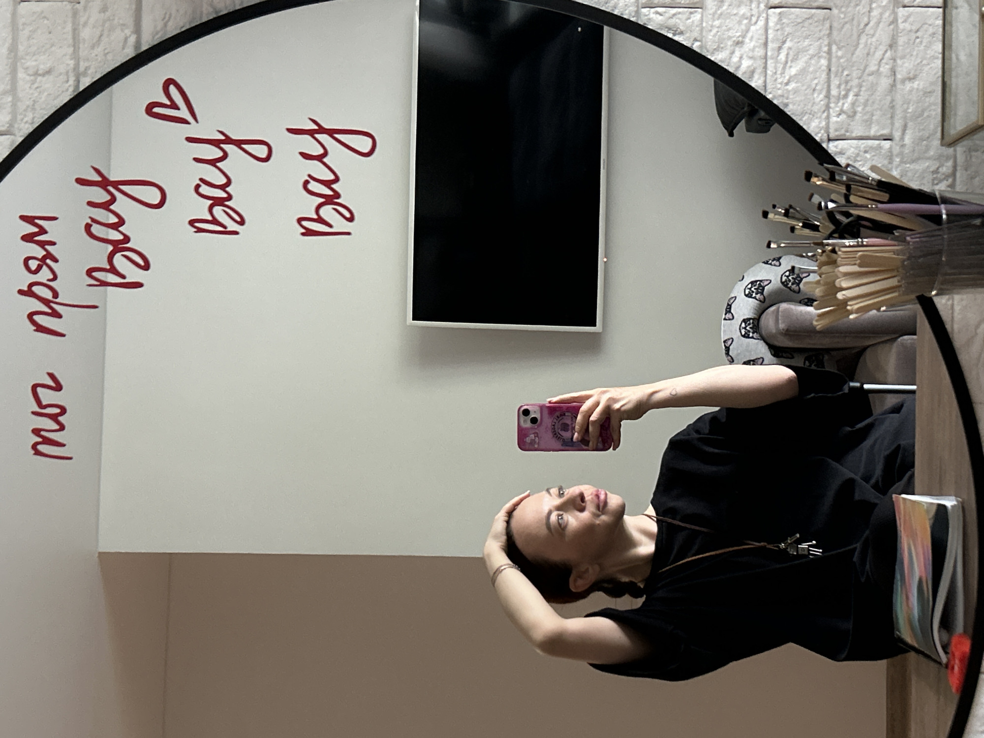 A mirrored image shows a woman holding a phone in a room. The room has a white wall with red writing on it, a television, and a collection of makeup brushes. The woman is wearing a black shirt and has her hair pulled back. The writing on the wall includes the word "Bay" repeated. The location may be in Chaikovsky, Russia.