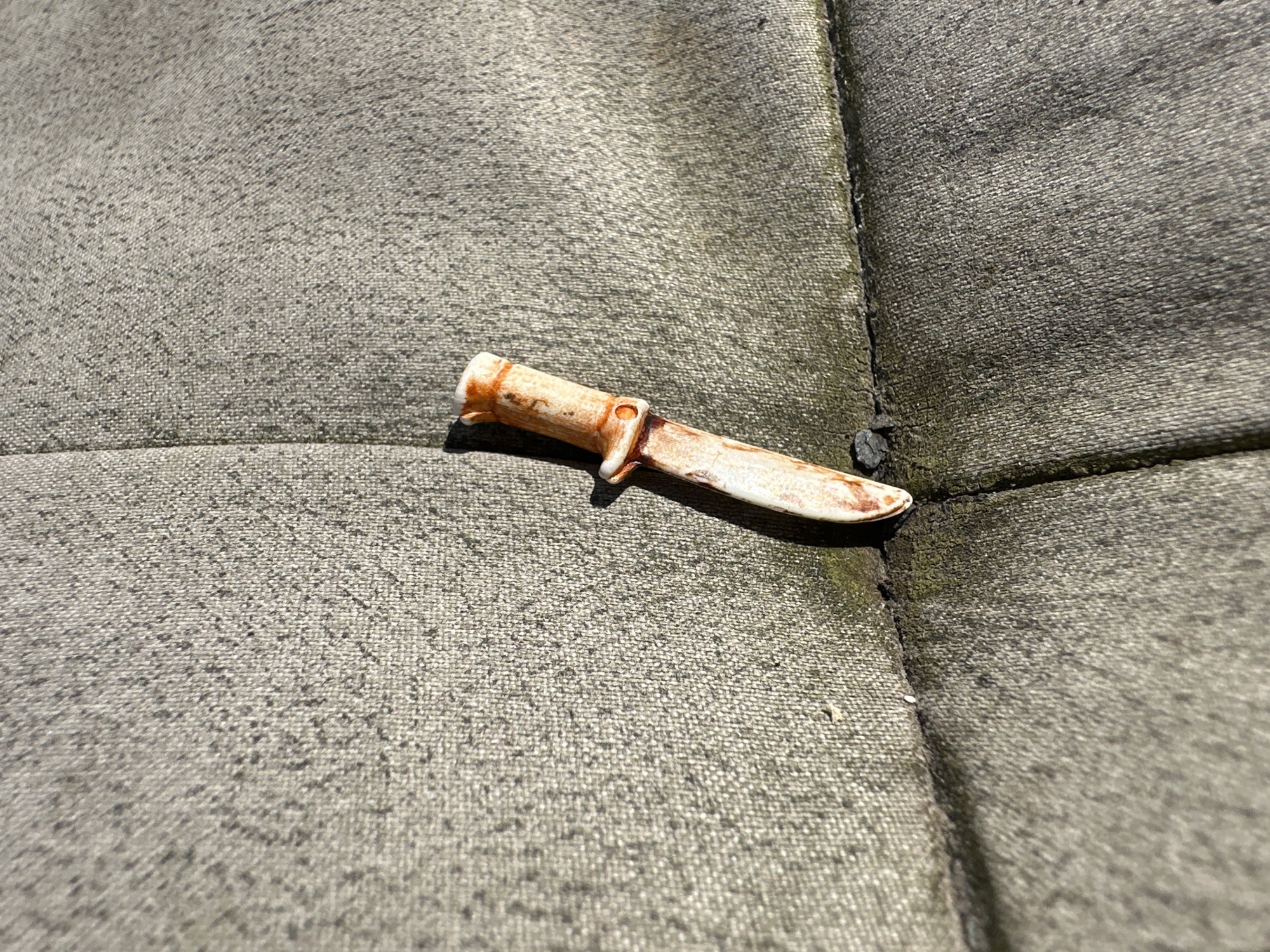 A small, toy-like knife with an orange-brown handle and a light-colored blade is resting on a textured, gray fabric surface. The fabric appears to be upholstery, possibly from a chair or couch, and shows signs of wear and possibly dirt or grime. There is a visible seam running vertically through the fabric to the left of the knife. The lighting suggests it is daytime, with shadows cast by the knife. The image was taken outdoors, as indicated by the natural lighting. The location is specified as Lelystad, Netherlands.