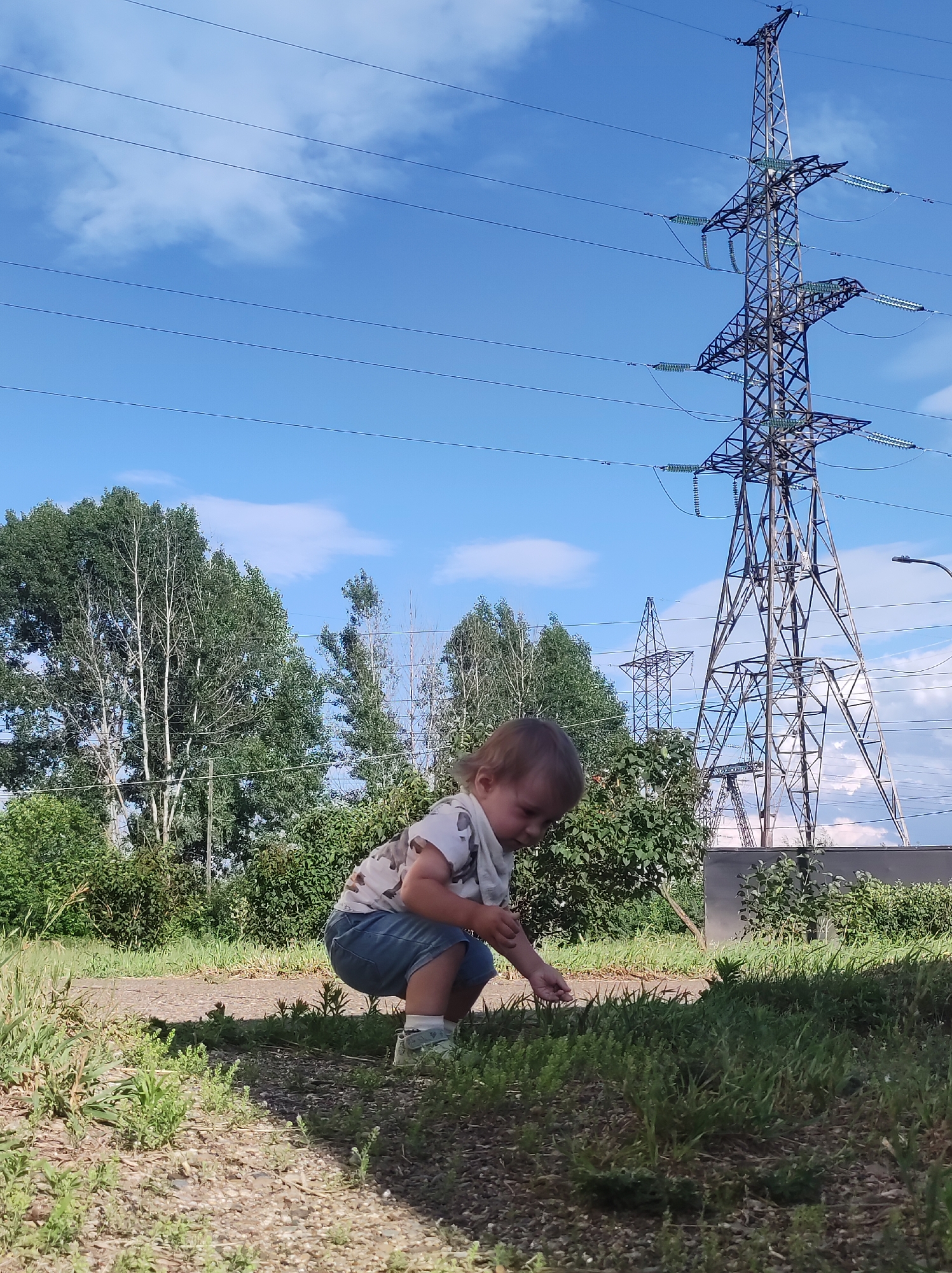 In the city of Chaikovsky, Russia, a young child, appearing to be a toddler, is captured in a crouched position on the ground, with their head bowed and hands near the grass, as if examining something. The child is wearing a t-shirt and denim shorts. The background of the image features a wide expanse of sky with scattered clouds, above a line of trees and a towering electricity pylon. Multiple power lines stretch across the sky, indicating an industrial or utility area within or near the city. The overall scene depicts a moment of childhood curiosity set against a backdrop of infrastructure and nature.
