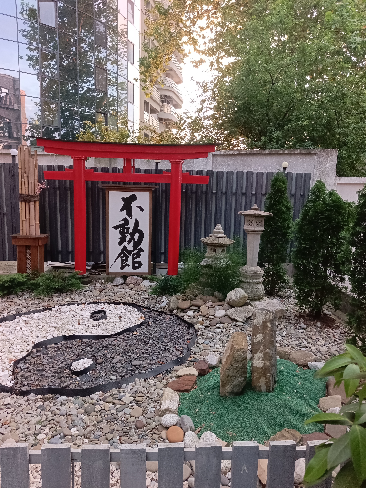 The image depicts a Japanese-style garden within an urban setting. A prominent red torii gate stands against a backdrop of a modern building with reflective windows and lush green trees. The garden features a yin and yang symbol created with white and dark gravel, surrounded by various rocks and stones. A traditional stone lantern and a smaller pagoda-like structure are visible, along with a vertical arrangement of bamboo poles. The ground is covered with a mix of pebbles and gravel, with a small patch of vibrant green turf in the foreground. The overall impression is one of tranquility and cultural fusion, juxtaposing Eastern aesthetics with an urban environment. The specific location of this garden in Sochi, Russia is not provided in the image.
