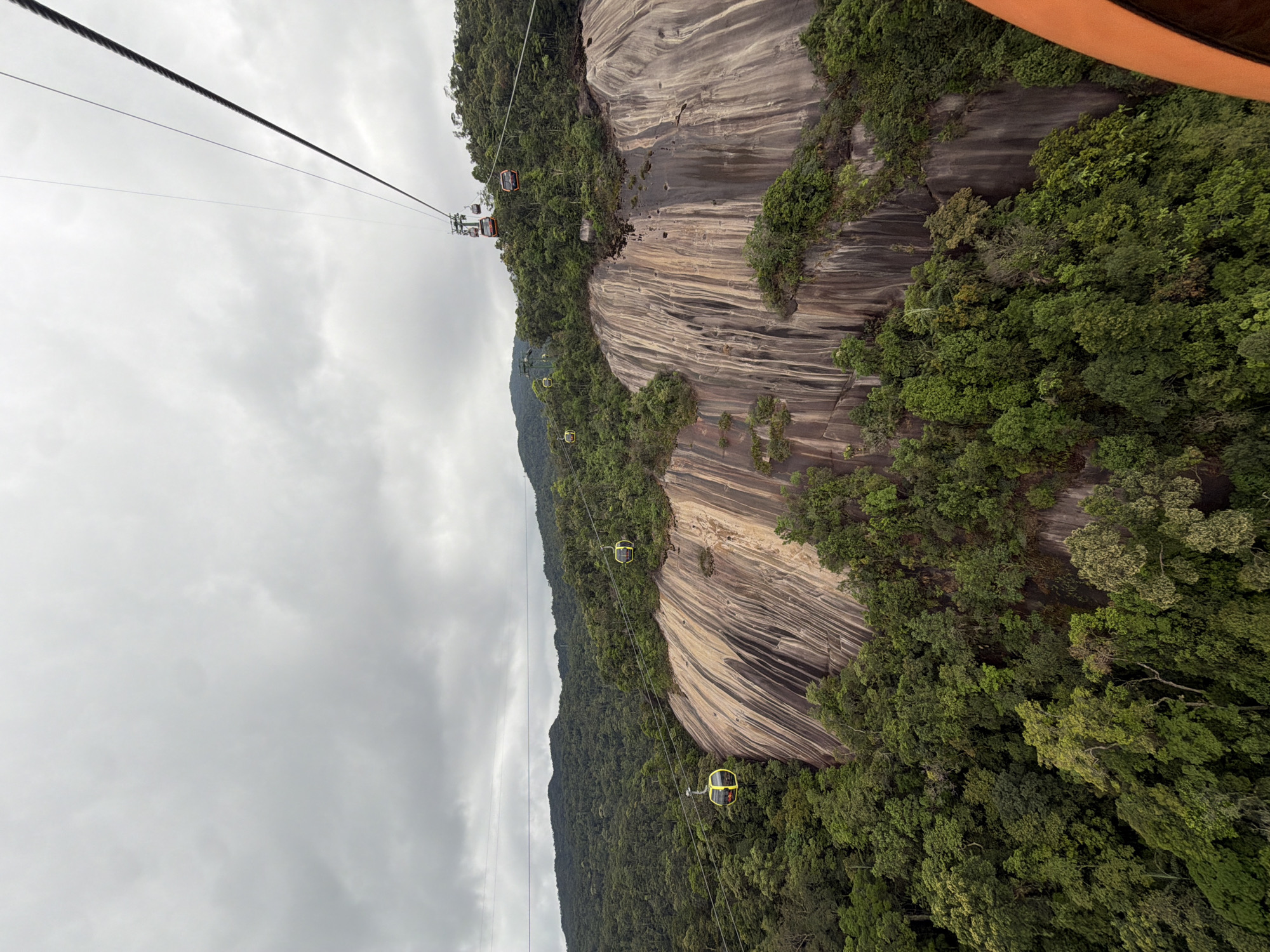 Captured from a moving gondola, the image presents an aerial view of the Bà Nà Hills mountain range in Đà Nẵng, Vietnam. The scene is dominated by a steep, striated rock face, heavily interspersed with lush green vegetation. Several gondolas, suspended by cables, are visible at varying distances, indicating the mountain's cable car system. Overcast skies create a diffused light, softening the contrast across the landscape.