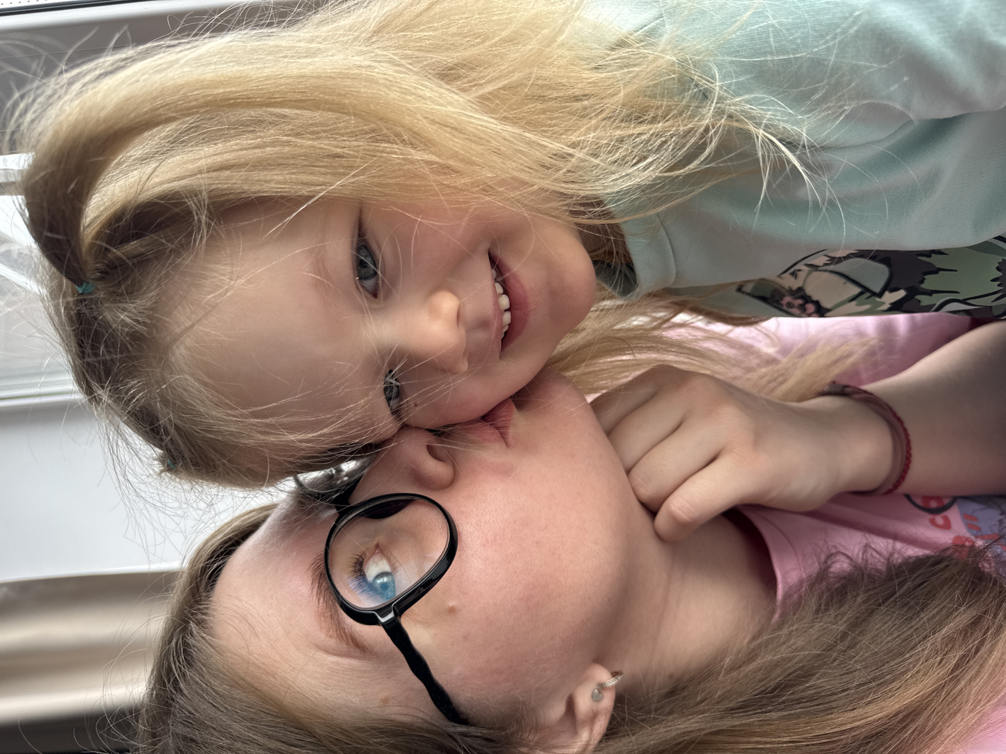 Two individuals are depicted in a close-up indoor portrait in Gornyi (Dvurechenskoe S/P), Russia. An adult female, positioned on the left, has light brown hair and wears black-framed circular glasses. Her left eye is visible, blue in color. She wears a pink t-shirt and has a small silver hoop earring in her left earlobe with an additional stud above it. Her right wrist (viewer's left) displays a thin red string bracelet. She is shown in the act of kissing the right cheek of a young blonde female child, who occupies the right side of the frame. The child has blonde hair, with a section secured by a light blue elastic band atop her head. She wears a light blue long-sleeved shirt with a partially visible abstract, camouflage-patterned graphic on the chest. The child faces the camera with a slight smile, revealing her upper teeth. The background is light-toned and indistinct, consistent with an indoor setting near a window or light source.