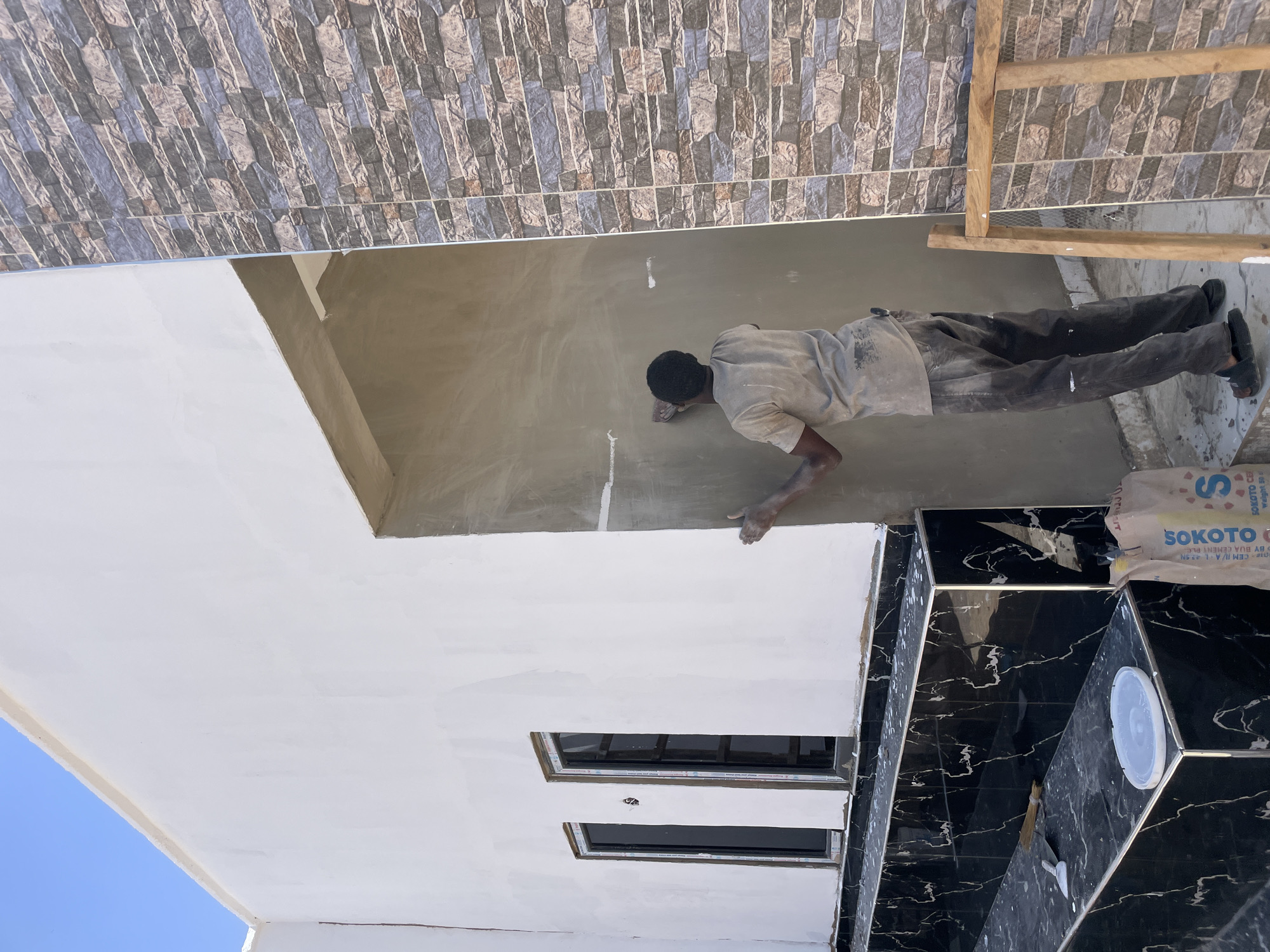 The image shows a construction scene, potentially in Jos, Nigeria, given the "SOKOTO CEM" cement bag present, as Sokoto Cement is a common cement brand in Nigeria. A man in a light-colored t-shirt and dark pants is smoothing the wall. A bag of Sokoto Cement is visible. The architecture includes brick or stone tiles on the exterior and white-painted surfaces on the building's underside. Black-and-white marble tiles are also visible in part of the construction. The sky is visible in the corner of the image. A window is also seen in the white painted surface. A wooden ladder is also visible.