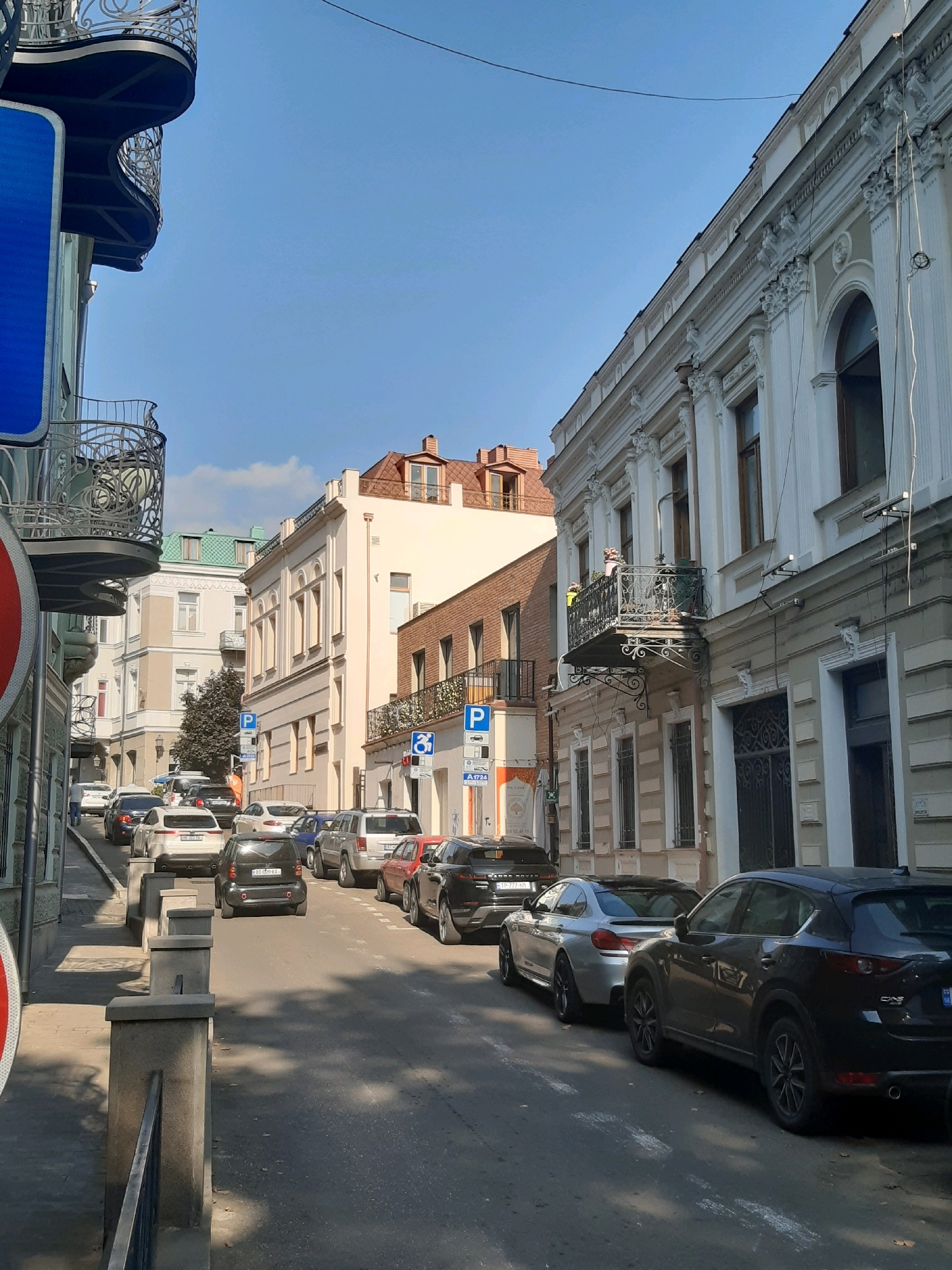 The image depicts a street scene in the city of Tbilisi, Georgia. A narrow street is lined with buildings featuring classic architectural details, such as ornate balconies and decorative facades. The street is filled with parked cars, including a black Mazda CX-5, a silver BMW, a black Smart car, and several other sedans and SUVs. A few people are visible, including a person in an orange vest standing near a parked car and another person walking on the sidewalk. The sky is clear blue with a few wispy clouds. The overall impression is of a typical day in a historic urban environment.
