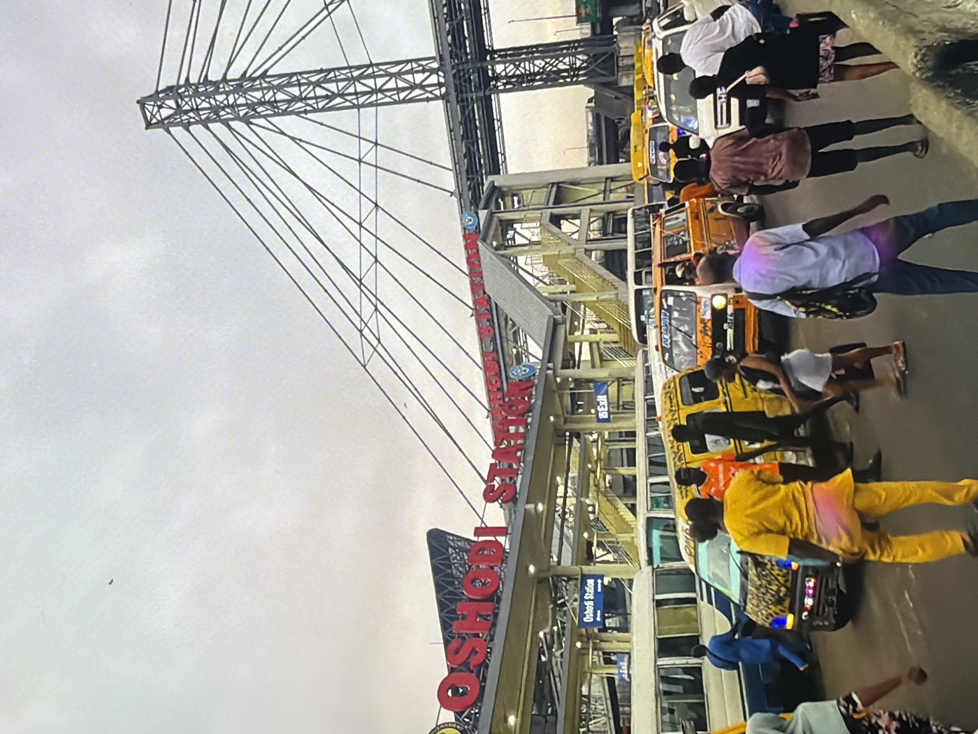 The image depicts a bustling scene at Oshodi Station, a transportation hub in Lagos, Nigeria. In the background, a prominent sign reads "OSODI STATION" in large red letters. Suspended above the station are cables of a large bridge or flyover, supported by a massive metal tower. Multiple buses and various colorful minibuses, commonly known as danfos, are visible, indicating active public transportation. People are seen walking, some seemingly engaged in commuting or interacting. The ground appears to be asphalt, typical of a busy urban environment. The sky is overcast, suggesting a cloudy or possibly rainy day. The overall impression is one of vibrant activity and a lively urban atmosphere.