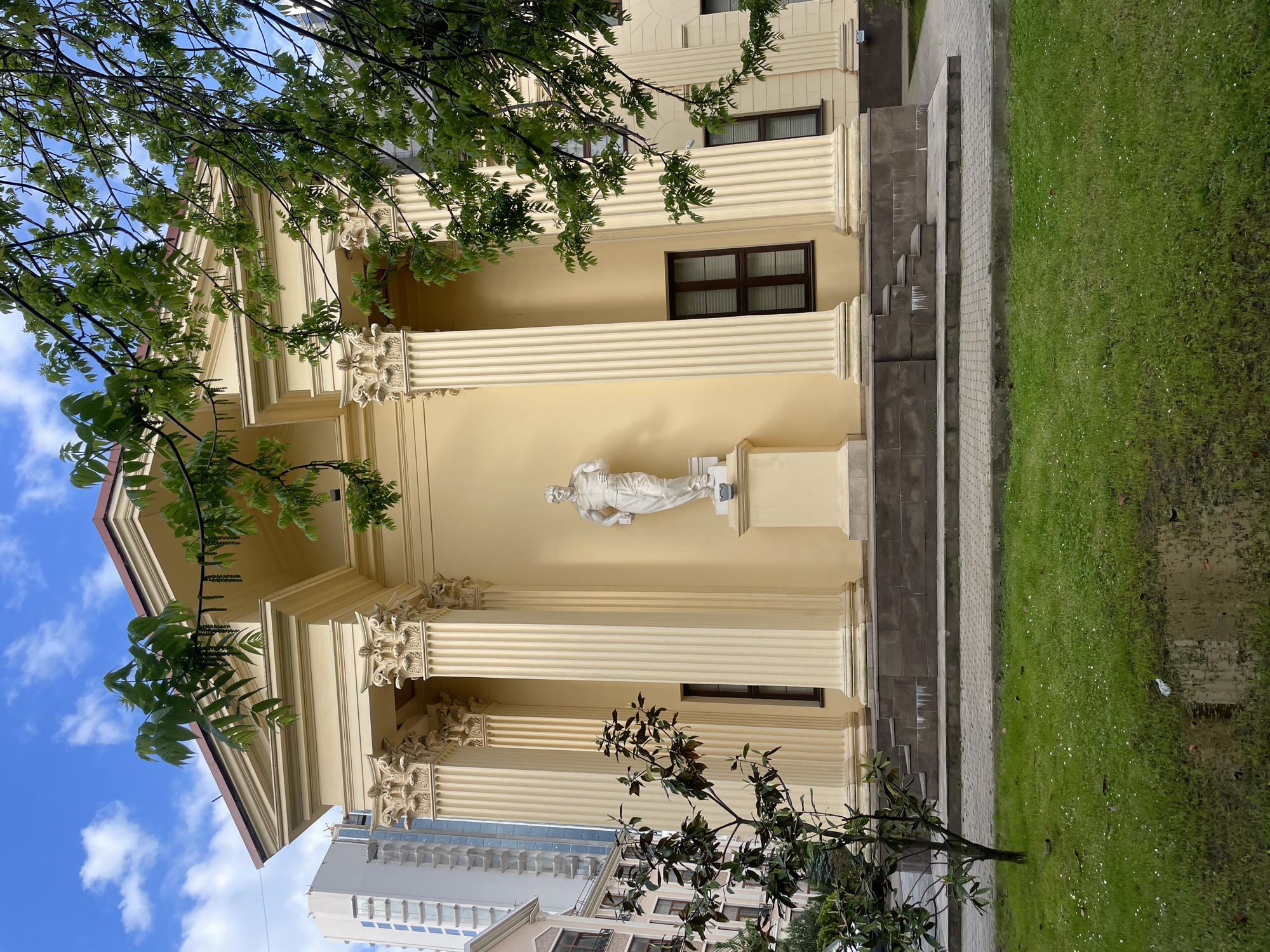 The image shows a portion of the Winter Theater in Sochi, Russia, viewed from below. The theater's facade is characterized by Corinthian columns, a pale yellow color scheme, and a classical pediment. A white statue is visible in an alcove on the facade. Trees partially obscure the building, with blue sky and clouds visible in the upper left corner. Part of a modern building can be seen at the bottom of the image. Grass and steps are adjacent to the building.