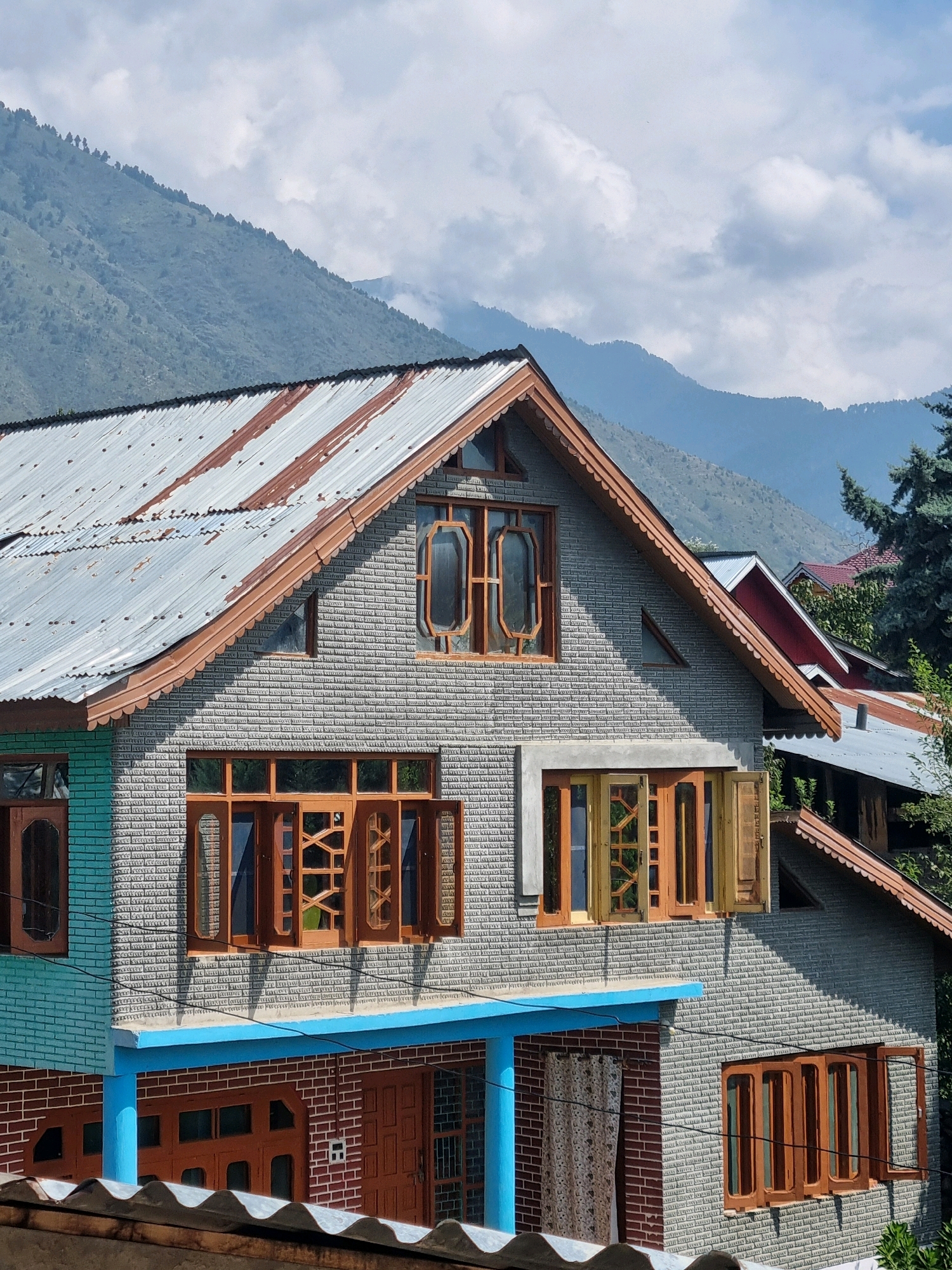 A residential building in Manigam, India, is captured in a photograph against a backdrop of misty mountains. The structure features a pitched, corrugated metal roof with visible rust. The walls of the house are constructed with grey bricks, intricately laid to create a textured pattern, and some sections are painted a light turquoise. Multiple wooden-framed windows are present, some of which are open, revealing their individual panes. The building has a vibrant blue horizontal trim running along its upper section. In the foreground, a portion of another corrugated metal roof is visible, suggesting a layered architectural design or proximity to other structures. The environment appears to be a rural or semi-rural setting, characterized by the presence of mountains and sparse vegetation, with a cloudy sky overhead. There are no people visible in the frame.