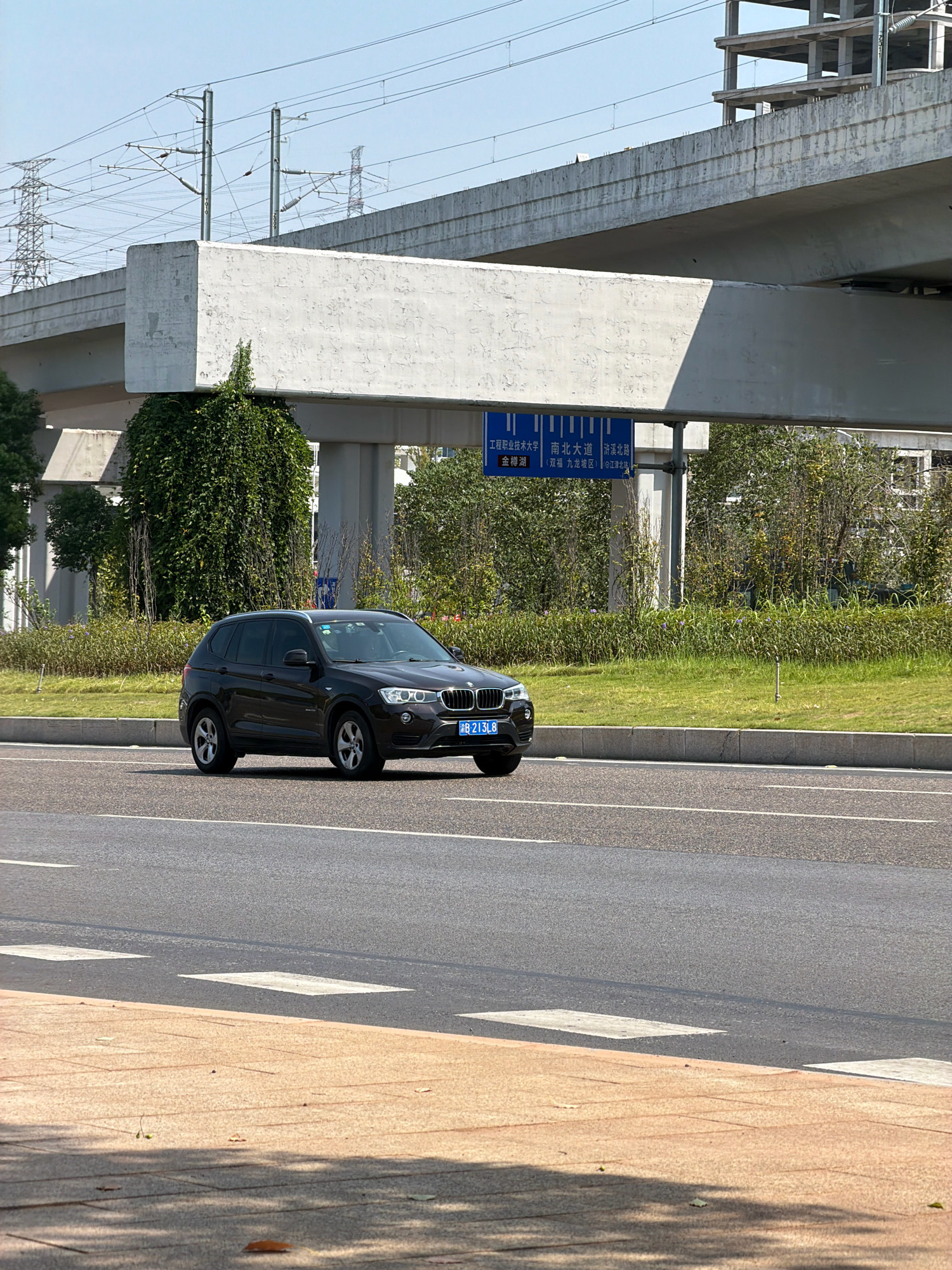 A dark-colored BMW X3 SUV is captured in motion on a paved road in Jiangjin, China. The vehicle's license plate reads "B 213L8". The road appears to be a multi-lane thoroughfare with white lane markings and a pedestrian crossing highlighted by white rectangular blocks. In the background, a large concrete overpass or bridge structure dominates the scene, with visible power lines and electrical towers extending across the sky. The environment is urban, with lush green grass and dense foliage bordering the road. A blue directional sign is affixed to the overpass structure, featuring Chinese characters and indicating destinations such as "Engineering Vocational and Technical University," "North-South Avenue," and "Zhixi North Road," along with specific location names like "Jinxi Lake" and "Jiulong District." The sky is clear and blue, suggesting fair weather. The image is a still photograph taken from a low vantage point, slightly to the right of the vehicle.