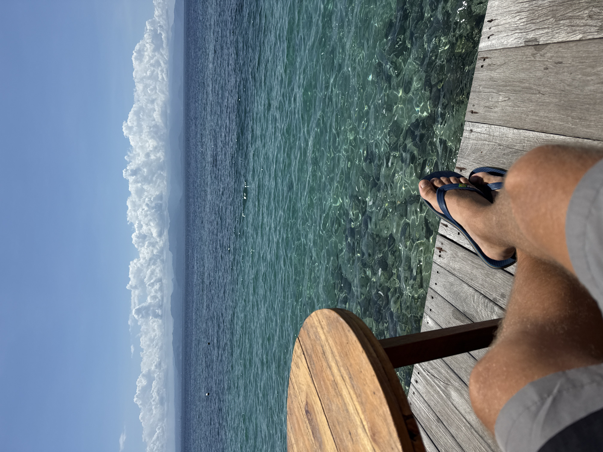 From a first-person perspective, the image captures a serene waterfront scene. A person wearing blue flip-flops is sitting on a wooden dock. The clear, turquoise water reveals the seabed beneath. To the left, the open ocean is separated from the sky by a line of heavy, white clouds. The location appears to be Nusa Penida, Indonesia.