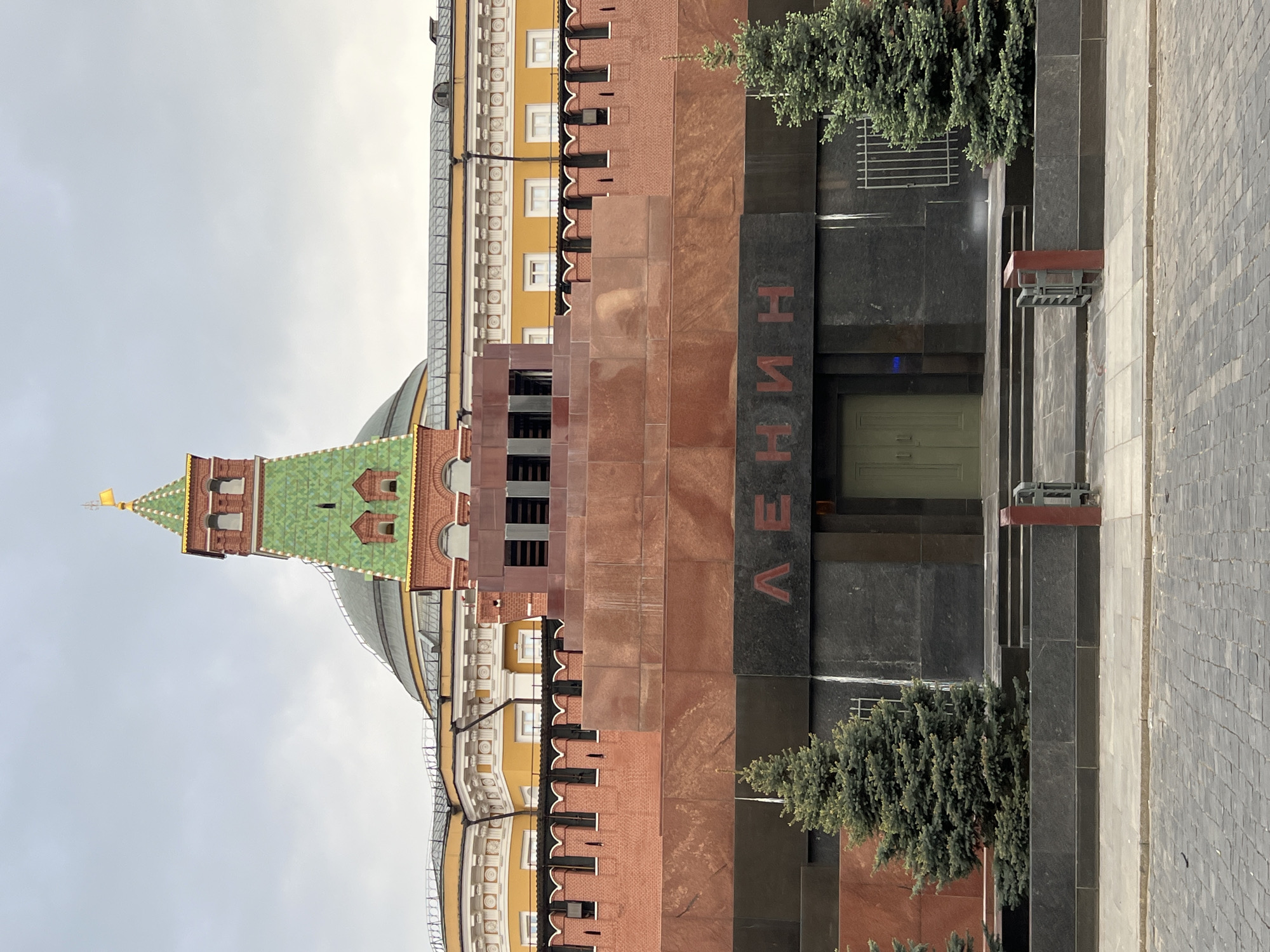 The image shows a partial view of Lenin's Mausoleum in Moscow, Russia. The building has a dark granite facade with the Cyrillic word "ЛЕНИН" ("Lenin") inscribed vertically. Two small evergreen trees flank the entrance. To the left of the mausoleum, a section of the Kremlin wall and one of its towers with a green roof are visible. A fragment of a yellow building is also visible. The ground in the foreground shows a paved surface. The sky is overcast.