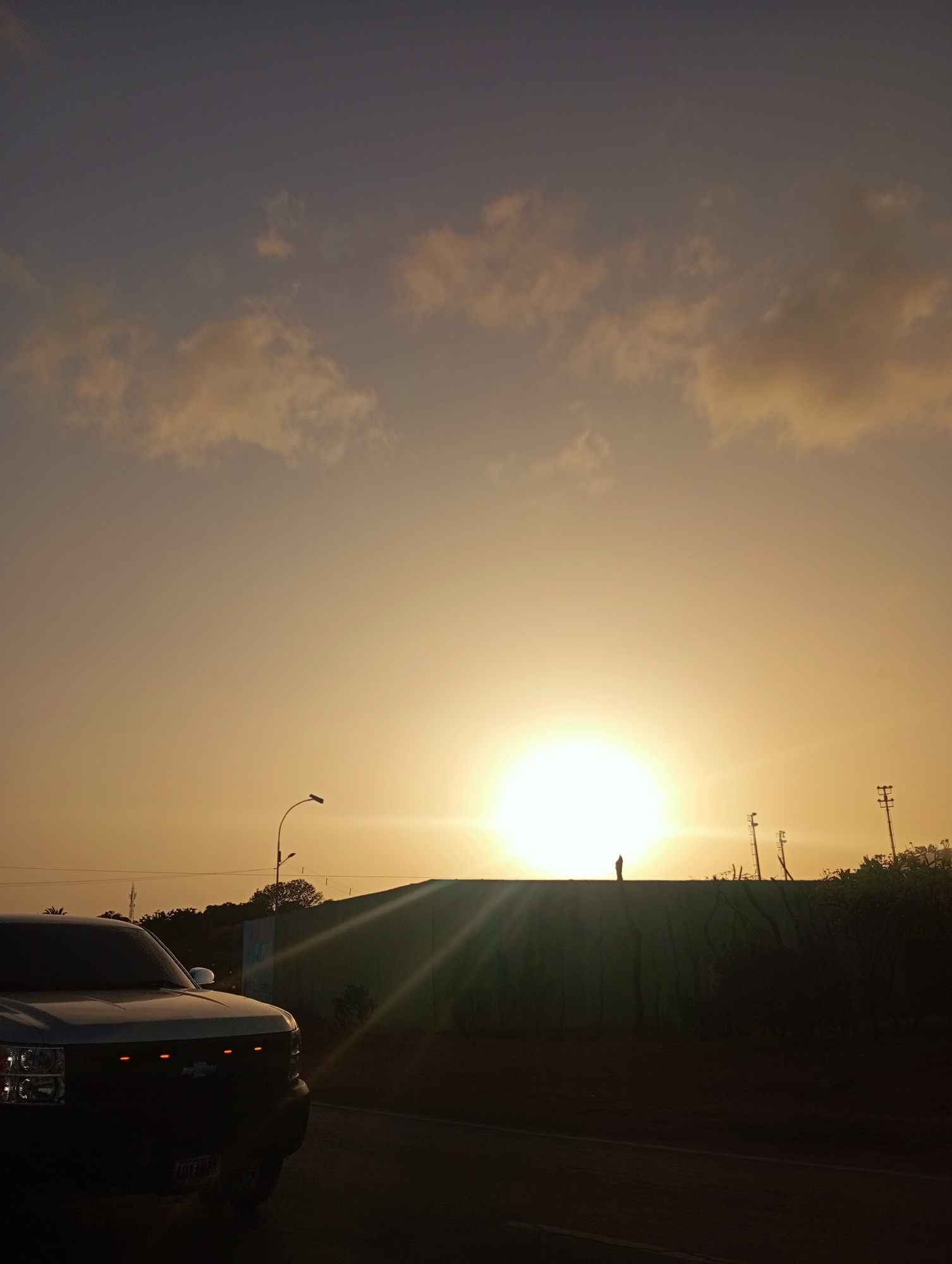 This photograph captures a breathtaking sunset or sunrise scene over a road in Juan Griego, Venezuela. The sky is the dominant feature, displaying a gradient of warm colors that transition from a darker, purplish-grey at the top to a brilliant orange and yellow near the horizon, where the sun is intensely bright and creates a noticeable lens flare. Scattered, light-colored clouds are visible, softly illuminated by the sun's glow.
In the foreground on the left, the front section of a dark-colored pickup truck is partially visible, angled towards the viewer. It appears to be a Chevrolet, identifiable by the general grille design and the characteristic orange marker lights integrated into the grille. The headlights and part of the hood are seen, with the vehicle appearing as a dark silhouette against the brighter background. A dark asphalt road occupies the bottom of the frame.
A long, dark wall or structure extends across the mid-ground, silhouetted against the vibrant sky and intense sunlight. A striking detail is a small, dark human figure standing atop this wall, positioned almost directly in front of the sun, creating a powerful silhouette. Behind the wall and to the right, several utility poles with cross-arms and dense foliage, including trees and bushes, are discernible as dark silhouettes. A single street light with a curved arm is also silhouetted against the sky on the left.
The scene conveys a sense of peaceful observation, likely at the end of the day. The strong backlighting from the low sun creates dramatic silhouettes and emphasizes the rich, warm colors of the sky. No legible text is visible in the image.