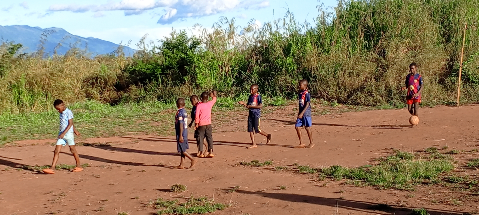 In Ngalula, Democratic Republic of the Congo, seven barefoot children stand on a dirt field bordered by high grass and shrubs. One child in a light blue collared shirt and light blue shorts walks left. Three children in the middle of the frame huddle together; one wears a pink long-sleeved shirt. Two children wear blue and red soccer jerseys and dark shorts. One child wears a blue and red soccer jersey and red shorts and balances a soccer ball on his right foot. Mountains are visible in the distance under a partly cloudy sky.