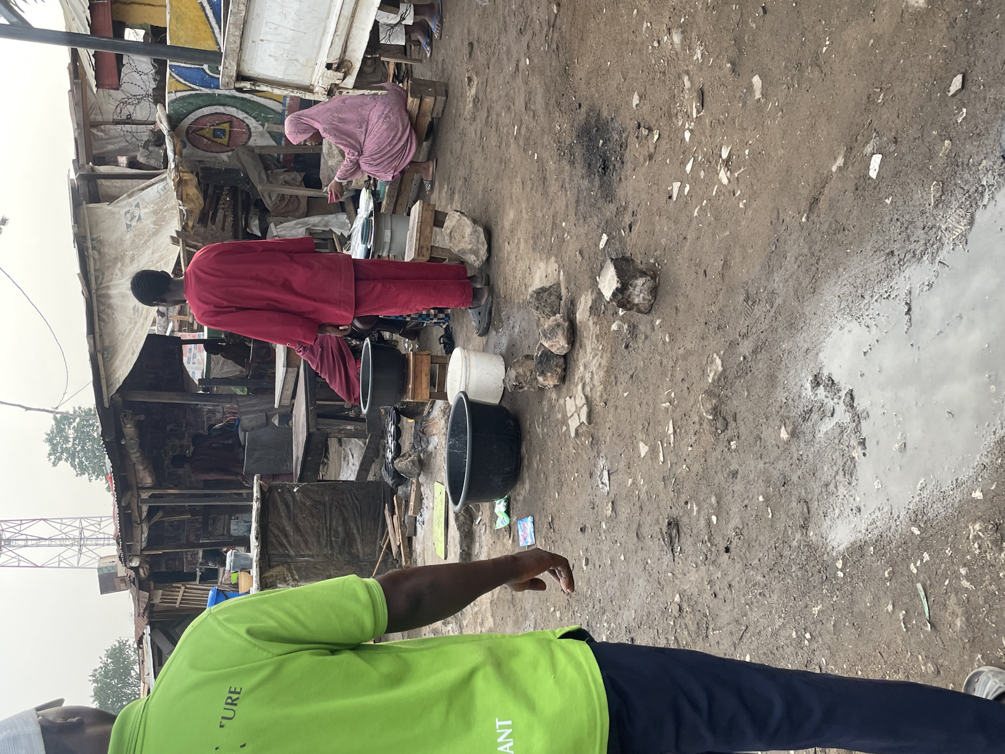 The image shows an informal settlement in Abuja, Nigeria. A man in a red robe stands near black and white buckets. A woman in a pink hijab sits on a wooden stool nearby. The ground is dirt, scattered with debris and puddles of water. A man wearing a green shirt with black pants stands in the foreground. A tower is visible in the distance. The settlement structures are built from makeshift materials.