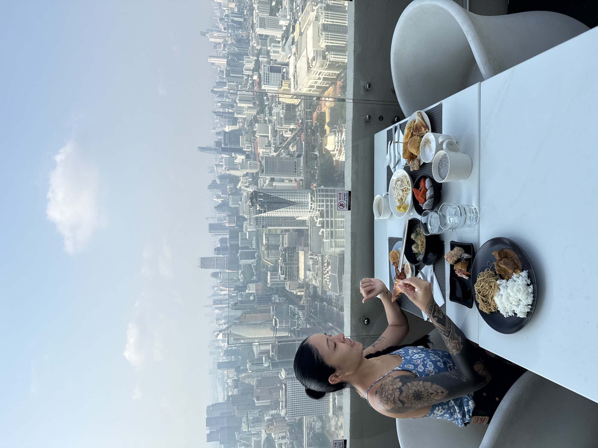 A woman is dining at a table with a panoramic view of Bangkok, Thailand. The table is set with various dishes, including rice, noodles, sushi, and sandwiches. The woman has tattoos on her arms and is wearing a blue floral dress. The city skyline is visible in the background, featuring a mix of high-rise buildings and lower structures under a hazy sky.