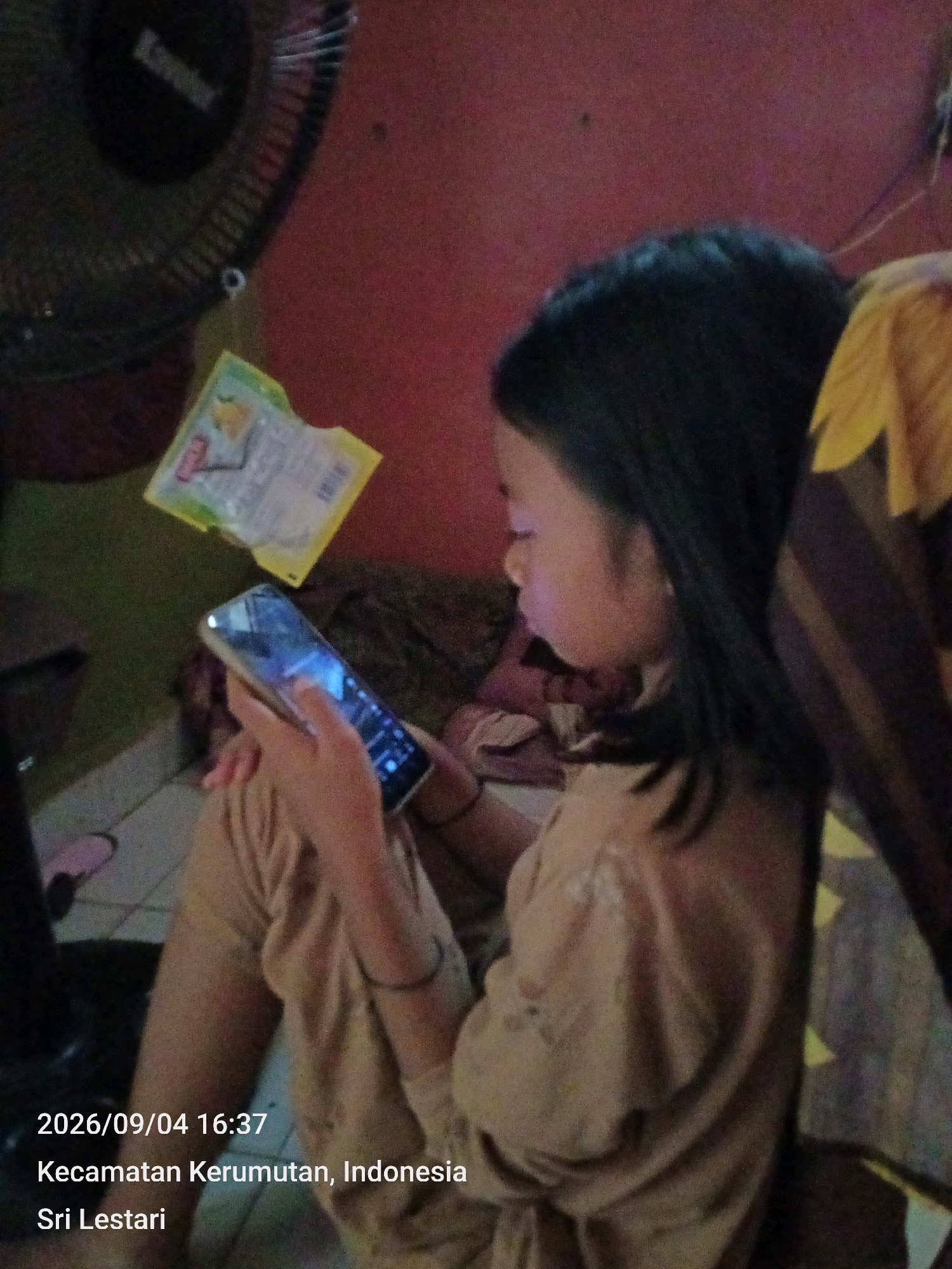 The image shows a young girl sitting on the floor, engrossed in her smartphone. She is wearing a light brown outfit. To her left, a black fan is partially visible. A yellow packet with some writing is hanging near the fan. The background wall is a reddish-orange color. The overall lighting is dim, suggesting an indoor setting. The bottom of the image has text that reads "2026/09/04 16:37 Kecamatan Kerumutan, Indonesia Sri Lestari". Based on the provided information, the location is Kecamatan Kerumutan, Indonesia.