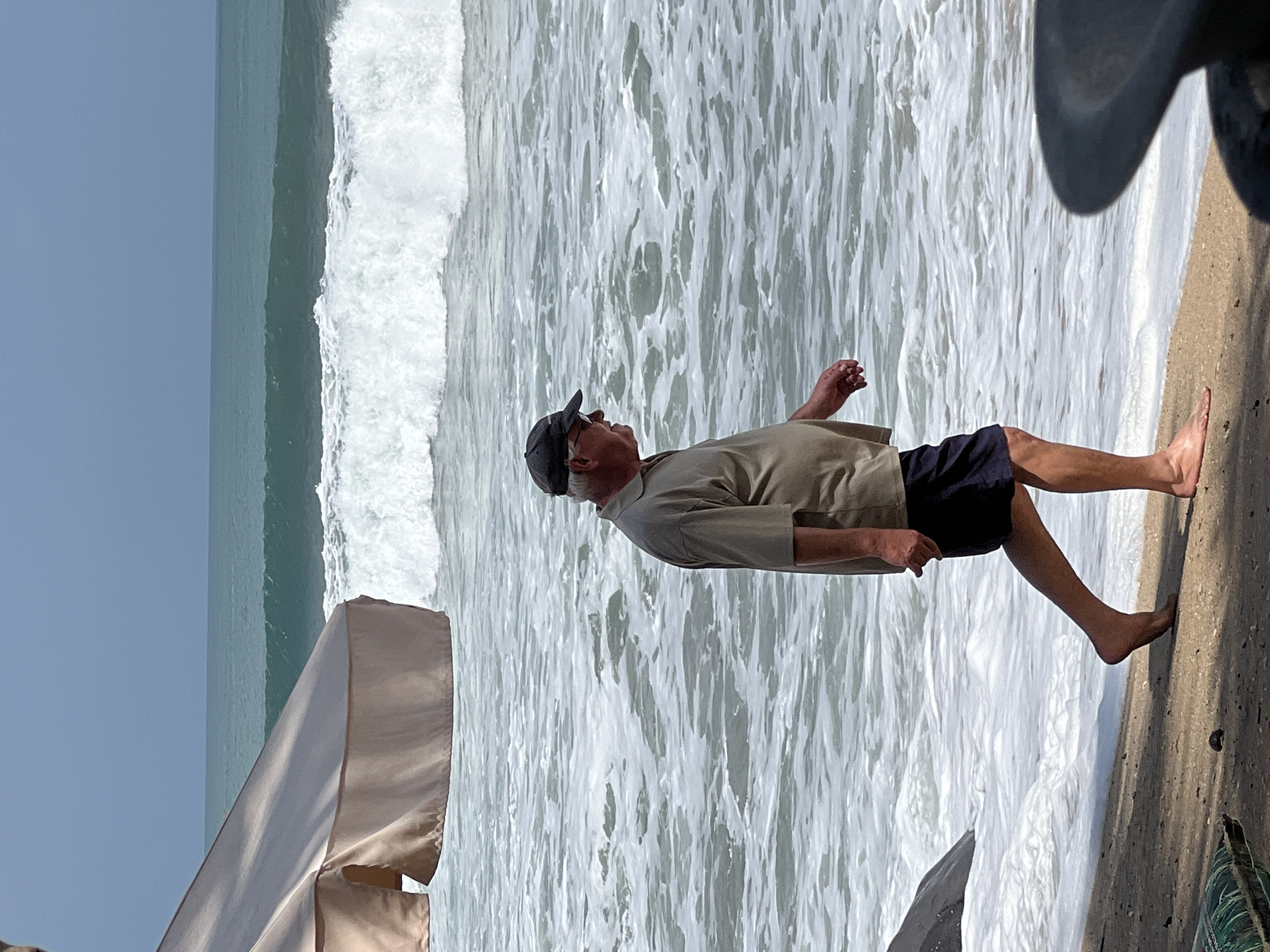An elderly man wearing a light brown short-sleeved polo shirt, dark blue shorts, and a dark baseball cap is walking along the sandy shore of Kuta Beach in Bali, Indonesia. The ocean is to his right, with moderate waves breaking and a foamy white surf reaching the shore. In the background, the sea stretches to the horizon under a pale blue sky. To the left of the frame, a portion of a large, beige beach umbrella is visible, casting a shadow. The man is in profile, facing towards the right of the image, with his hands clasped loosely in front of him. His left leg is extended forward, mid-stride, and his right foot is on the sand. The sand in the foreground has some texture and debris, with a dark object, possibly a table or chair leg, visible in the lower right corner, partially obscuring the view.