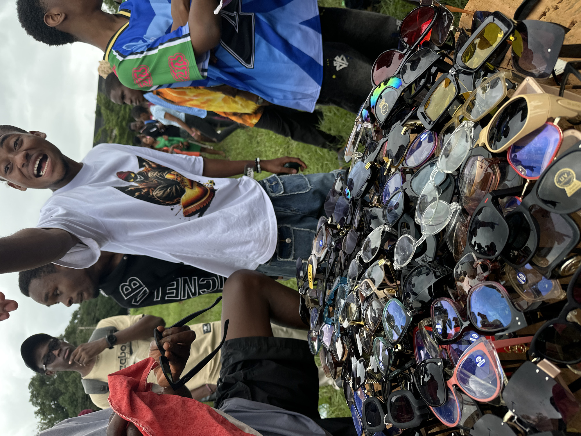 In Ihiagwa, Nigeria, a group of young people are gathered, with a table laden with sunglasses in the foreground. The individuals are dressed casually, with one person wearing a white t-shirt featuring a graphic design, another in a black hoodie with white lettering, and a third in a blue and green athletic jersey. A red cloth is held by a person on the left side of the frame, near a pile of sunglasses. The background consists of green grass and trees, suggesting an outdoor setting, possibly a market or community gathering. The sky is overcast, indicating a cloudy day. The sunglasses on the table display a variety of styles, colors, and lens tints, suggesting they are for sale. The overall atmosphere appears to be informal and social.