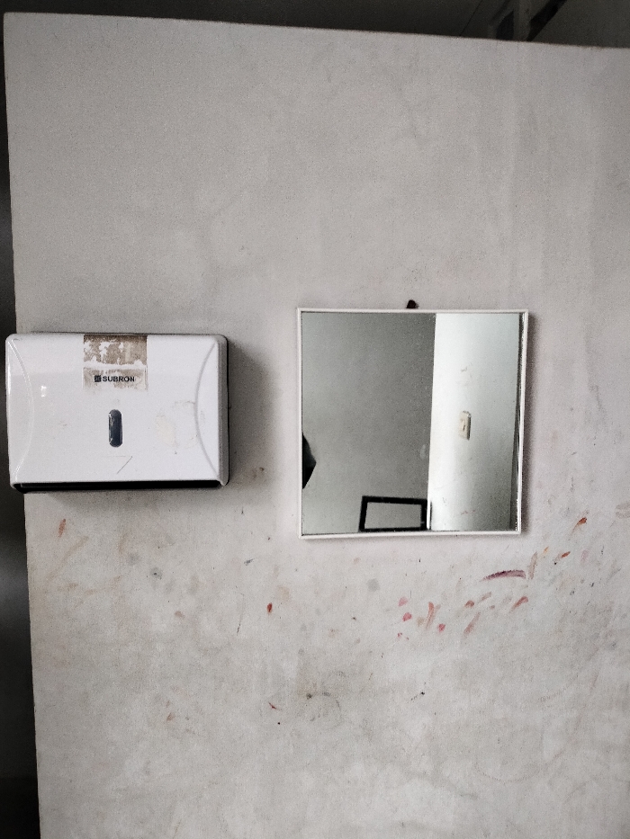 The image displays a light-colored, textured wall with a paper towel dispenser and a square mirror mounted on it. The wall, possibly concrete or painted plaster, shows considerable wear, characterized by numerous smudges, dark marks, and reddish splatters, suggesting a utilitarian or well-used environment.
On the left, a white, rectangular dispenser, likely for paper towels or wipes, is affixed to the wall. It features a visible dark slot. A label on its front is partially peeled and worn, but the brand name "SUBRON" is discernible, accompanied by a small, possibly registered trademark, symbol.
To the right of the dispenser, a square mirror with a simple white frame hangs on the wall. The mirror reflects portions of an interior space. Visible in the reflection are a dark, indistinct silhouette on the left, a rectangular frame (possibly part of a window or furniture) towards the bottom-center, and a bright white vertical structure, perhaps a wall or pillar, on the right side of the reflection. The reflections imply a simple, possibly industrial or functional indoor area.
No specific activity is occurring; the items are stationary fixtures. The overall scene suggests an indoor location, possibly a public restroom, workshop, or utility area, in Dramaga, Indonesia. There are no explicit cues for the time of day, but the lighting is consistent with an indoor setting.