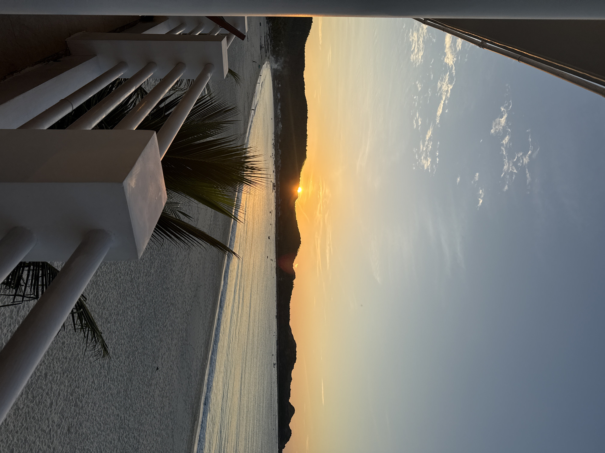 A serene beach scene unfolds at dusk, captured from an elevated vantage point, likely a balcony or veranda. The foreground is dominated by a white, modern railing, its clean lines contrasting with the natural elements beyond. A tall, white pillar stands to the left, part of the structure from which the photo is taken, casting a shadow on the lower foreground. Palm fronds intrude into the frame from the right and center, their dark green leaves adding a touch of tropical lushness.
The middle ground reveals a wide expanse of pale, sandy beach, gently sloping towards the calm sea. The sand is textured, showing subtle undulations and a scattering of what appear to be small shells or debris. A few distant figures can be seen walking along the shoreline, mere specks against the vastness of the beach. Along the edge of the water, a gentle wave washes ashore, leaving a glistening wet line on the sand.
In the distance, a crescent-shaped bay is embraced by dark, undulating hills. The setting sun, a vibrant orb of orange and yellow, is partially obscured by the crest of one of these hills, casting a warm, golden glow across the scene. The sky above is a gradient of soft blues and oranges, with wispy white clouds scattered across it. Several small boats are visible on the water, silhouetted against the radiant sunset. The overall impression is one of tranquility and natural beauty, with human presence minimal and unobtrusive.