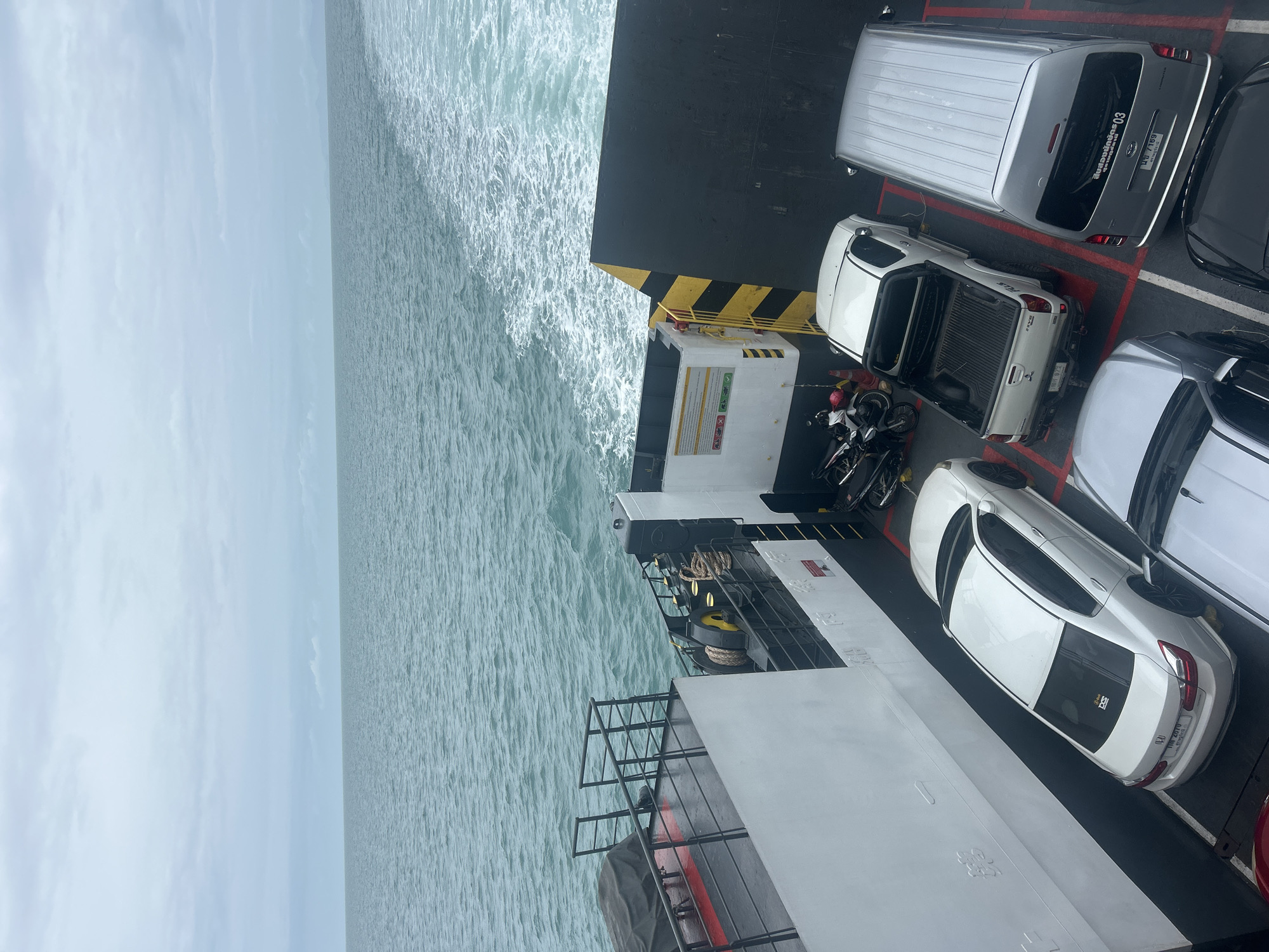 The image is a high-angle, full shot taken from a ferry on the ocean, likely in Thailand. The ferry is carrying several vehicles, including a white sedan, a white pickup truck, and a silver van. The vehicles are parked in designated lanes marked with red and white lines. In the background, the ocean is visible with a wake trailing behind the ferry. The sky is overcast, and the overall lighting is muted. There are no people clearly visible in the image, but there is a motorcycle parked on the ferry deck, along with some safety cones and equipment. The side of the ferry, with its white exterior and black and yellow striped railing, is also visible.