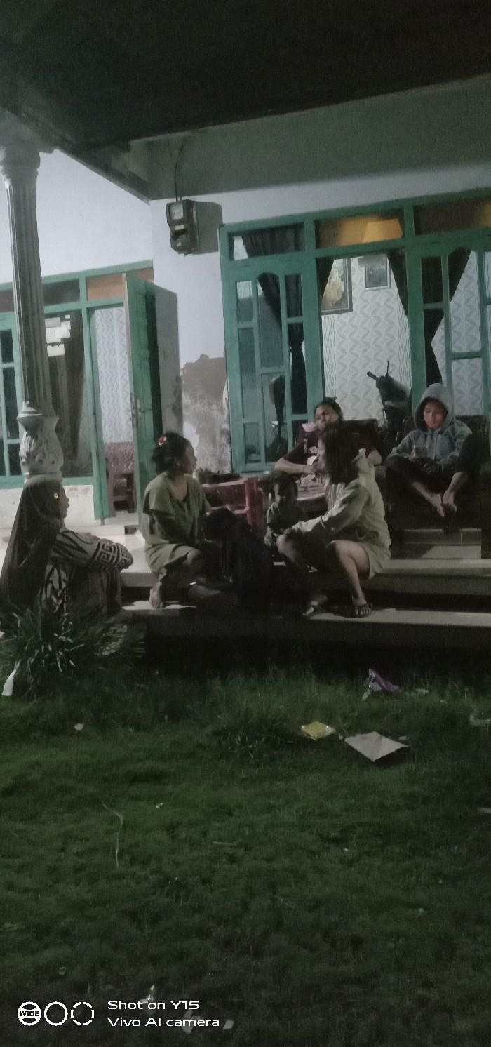 An outdoor scene captured at night or dusk in Ranugedang, Indonesia, depicts a group of approximately six individuals gathered on the steps of a building. The building features light-colored walls, teal-green window and door frames, and a fluted white column on the left. An electrical meter box is mounted on the wall above the windows. Through the windows, patterned curtains and framed artwork are visible. The foreground consists of dark green grass with several pieces of discarded litter, including a yellow wrapper, a purple wrapper, and a flat white item.
The individuals are seated on various steps. On the far left, a person wearing a dark head covering and a striped garment is seated low, facing right. Adjacent to them, a person in a light green top is seated on a step. Towards the center, a child and other adults are partially obscured. On a higher step, a person in a light-colored hooded sweatshirt and shorts sits with bare feet. On the far right, another person in a grey hooded sweatshirt is seated, holding a clear glass or cup. A dark-colored motorcycle or scooter is partially visible behind the group, near the building entrance. The individuals appear to be engaged in social interaction.