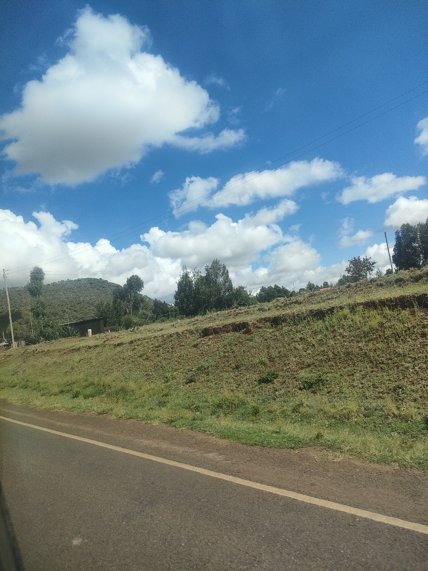 The image shows a rural landscape in Kisima ward, Kenya. A paved road with a yellow dividing line stretches across the foreground. Adjacent to the road, a grassy embankment rises, leading to a plateau with scattered trees and sparse vegetation. A building is visible among the trees. In the background, a hill is covered with trees. The sky is bright blue with scattered puffy white clouds. Overhead power lines are visible.
