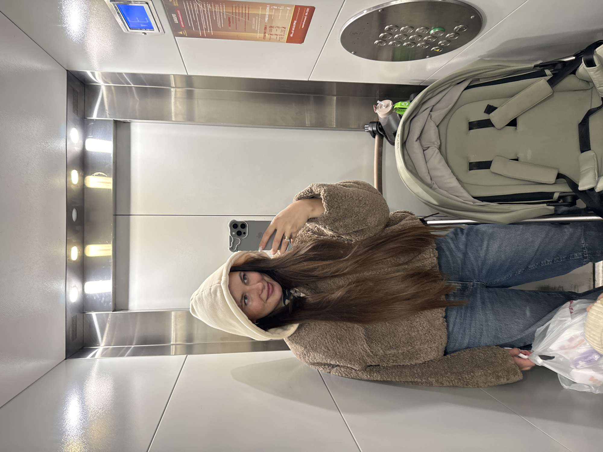 The image shows a woman taking a selfie inside a modern elevator, likely in Odintsovo, Russia. The woman, with long brown hair, is wearing a light brown hooded fleece jacket and blue jeans. She holds a grey smartphone to capture the moment and is smiling faintly at the camera. A light-colored baby stroller with a padded interior and safety straps is positioned next to her, indicating she is traveling with an infant, though the child is not visible. A white plastic bag with contents is seen near her feet.
The elevator interior is clean and brightly lit, featuring light-colored walls and floor with reflective surfaces. Vertical strips of light illuminate the side of the elevator. The control panel is located at the top of the elevator, displaying numerous circular buttons and a digital indicator showing "11" or "F1," likely denoting the current floor.
Above the control panel, an information placard with Cyrillic text is visible. It includes a red banner with "ВАЖНО!" (IMPORTANT!) and further details such as "КРУГЛОСУТОЧНО" (24/7), "ОБСЛУЖИВАНИЕ" (SERVICE), and a phone number "94-000," confirming the Russian location. The scene depicts a casual, everyday activity.