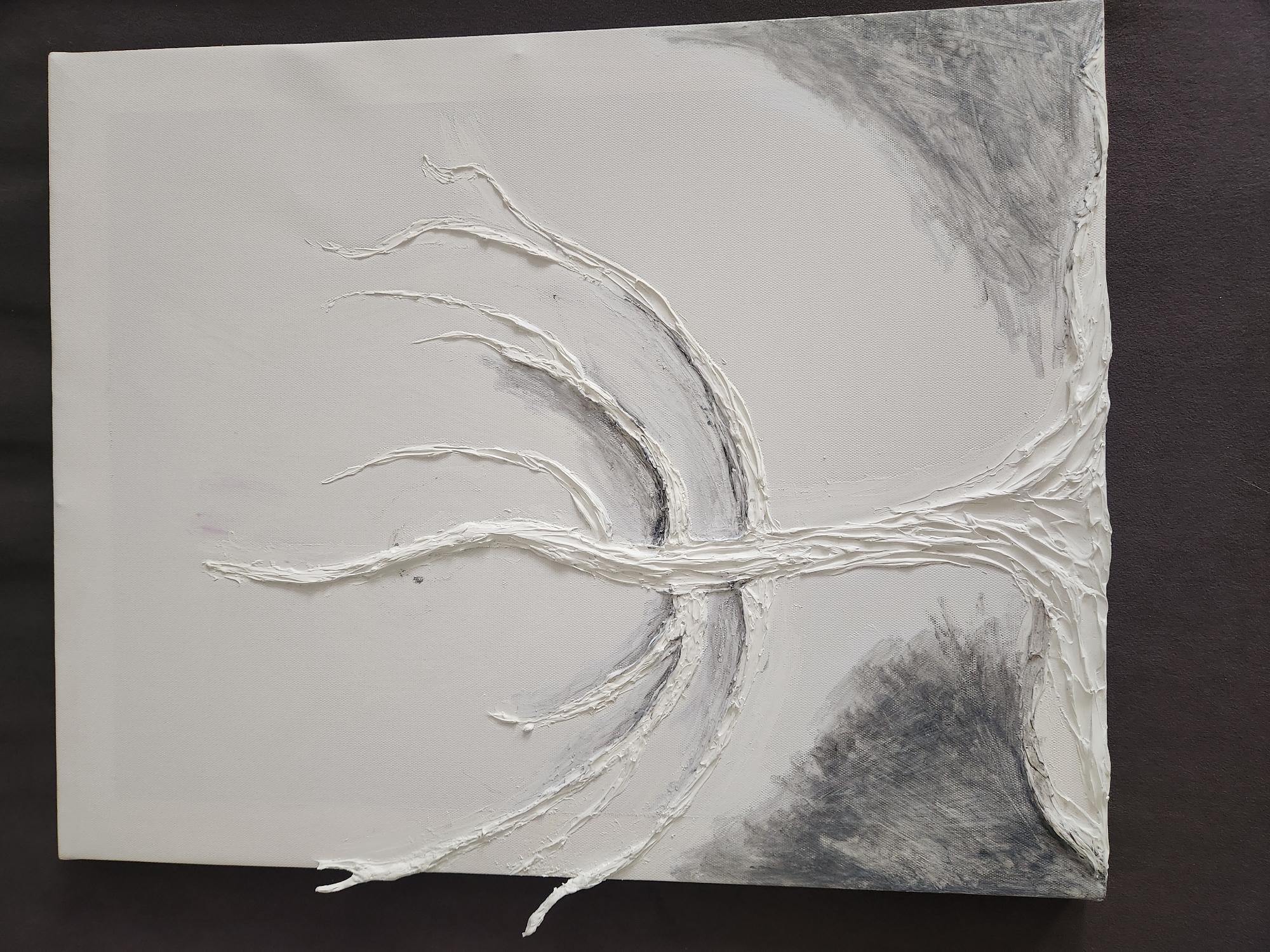 The image displays a close-up, overhead view of a textured painting on canvas. The artwork features a stark white, relief sculpture of a bare tree with gnarled branches. The tree's base and trunk are rendered with thick, impasto-like applications of white paint, creating a sense of ruggedness. The branches extend outwards across the canvas, thinning as they reach the edges. Some of the branches have small, twig-like extensions. The background is a subtle blend of off-white and grey, with darker, smudged grey areas suggesting foliage or ground cover on the left and right sides of the tree. The texture of the canvas is visible beneath the paint. The painting is presented against a dark grey, textured surface, likely a table or backdrop. The scene depicted is an artwork, and its location within the city of Vniissok, Russia, is not discernible from the image itself. There are no people, actions, or interactions visible.