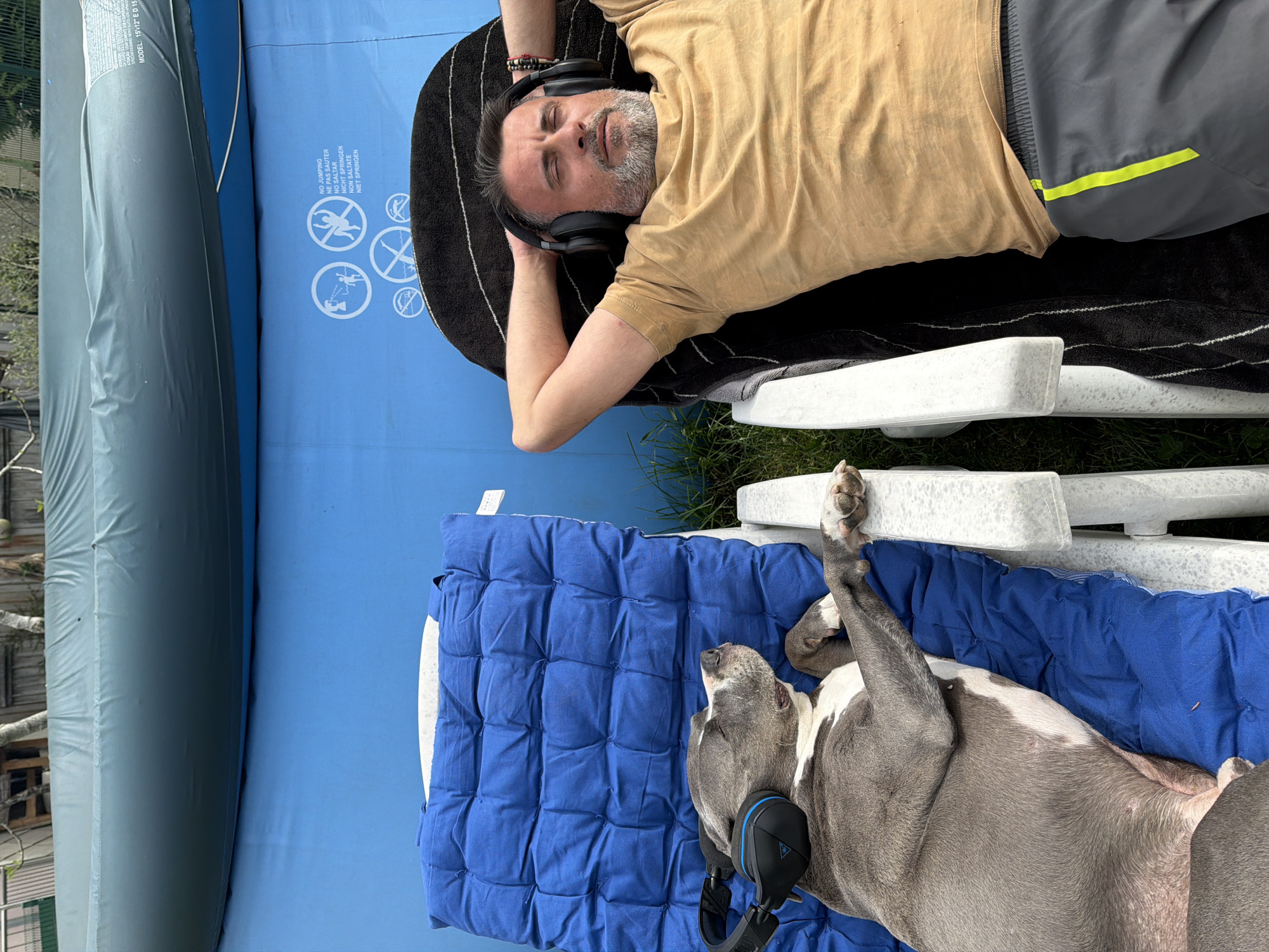 A man and a dog are relaxing outdoors. The man, wearing headphones, a beige t-shirt, and gray shorts with a yellow stripe, lies on a black towel atop a white lounge chair. The dog, a gray and white pitbull, also wears headphones and rests on a blue quilted cushion atop another white lounge chair. A blue inflatable pool surrounds the chairs. A gray inflatable tube is next to the pool. The location is in Esch-sur-Alzette, Luxembourg.