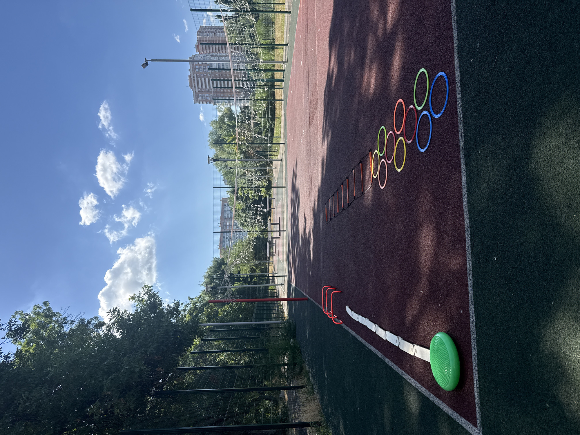 This is a daytime, eye-level, full-shot photograph of an outdoor sports court in the city of Krasnodar, Russia. The court is marked with red and dark green surfaces, separated by white lines. In the foreground, a green balance disc is placed on the court. To the left, there are several red hurdles lined up, and a white marker line extends from them. Further into the court, a speed ladder is laid out, followed by a series of colorful hoops arranged in a curved line. In the background, a volleyball net is set up across the court. Beyond the court, trees, a fence, and modern apartment buildings are visible. The sky is blue with scattered white clouds. There are no people in the image.