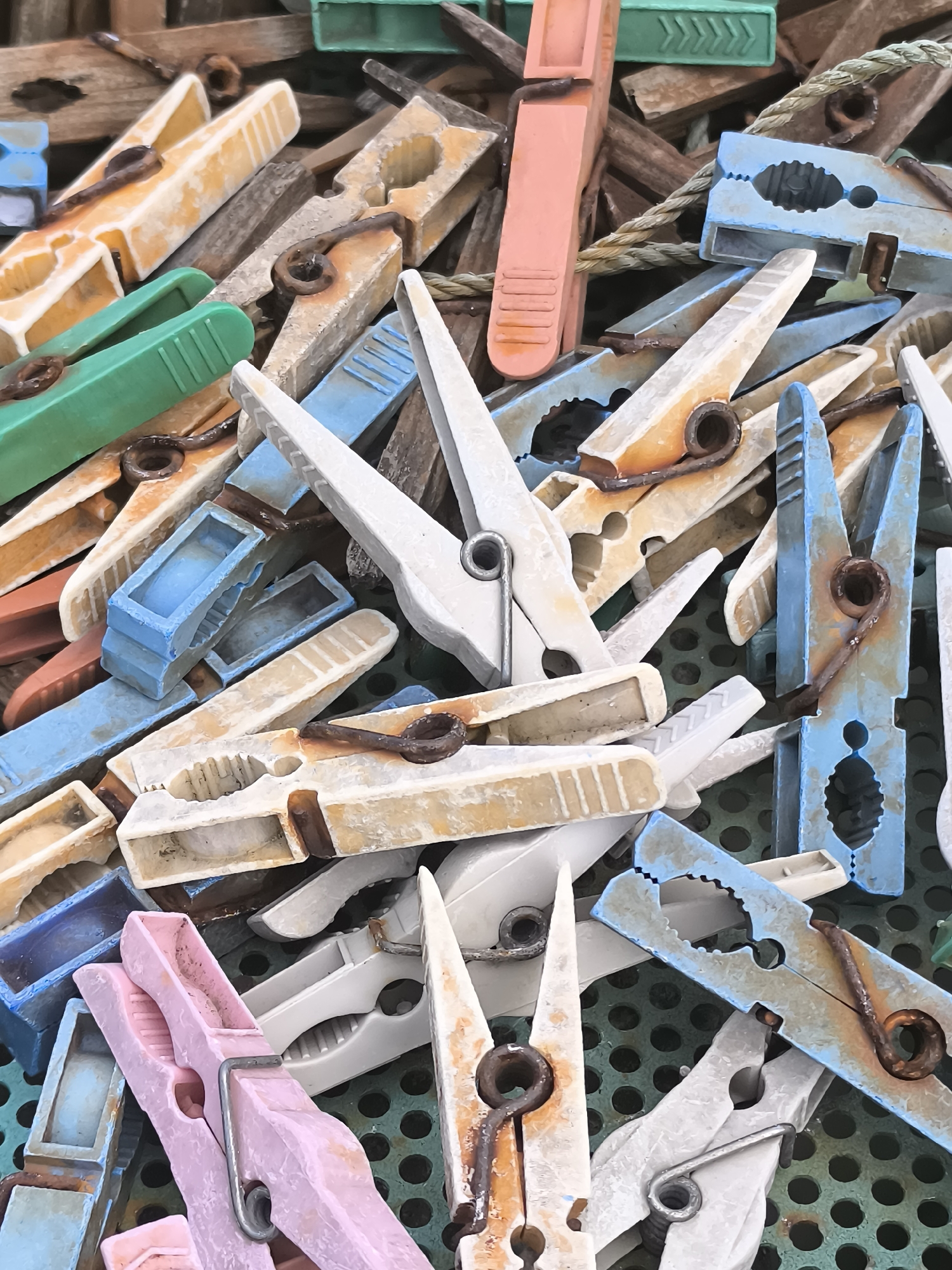 This is a close-up, overhead shot of a large, disordered pile of old, weathered clothespins. The clothespins are made of plastic and are various colors, including faded pink, light blue, cream, orange, and green. Many of them show significant rust on their metal springs and are covered in dust and grime, indicating they have been used extensively and exposed to the elements. The clothespins are jumbled together on what appears to be a green perforated surface, possibly a drying rack or a similar outdoor utility item. In the background, there are glimpses of what might be wooden planks and rough rope, suggesting an outdoor setting, possibly a backyard or shed. The lighting is natural and diffuse, with no strong shadows. The image conveys a sense of disarray and neglect, with the once functional items now appearing worn and abandoned. The location of Dobrogea Veche, Moldova is not visually represented in the image itself; this detail is provided as external context.