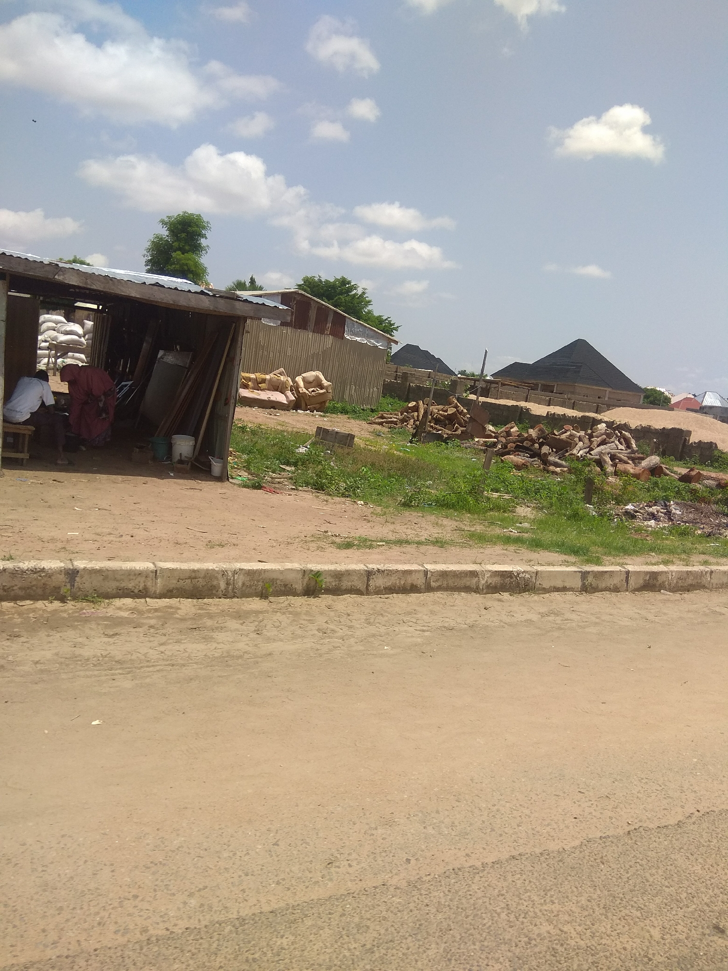 This image captures an outdoor scene in Jalingo, Nigeria, under a bright, partly cloudy sky, suggesting it is daytime. The foreground features a dusty, unpaved area with a concrete curb, likely a roadside.
Dominating the middle ground is a makeshift structure with a corrugated metal roof, resembling a small shop or shelter. Inside, two individuals are visible: one person in a white shirt sits on a wooden stool, and another, possibly a woman, is wearing a maroon garment. Stacks of white sacks, likely containing goods, are visible behind them. Various objects, including planks of wood and buckets, are also present within and around the shelter, indicating a commercial or storage area.
To the right of the shelter, a collection of cut logs lies on the ground, suggesting a wood supply or construction site. In the background, several buildings with dark, pyramidal roofs are under construction or recently completed, along with other structures that appear to be houses. The overall setting suggests a developing area with ongoing construction and commerce. The scene appears active, with people and materials present. There is no visible text in the image.