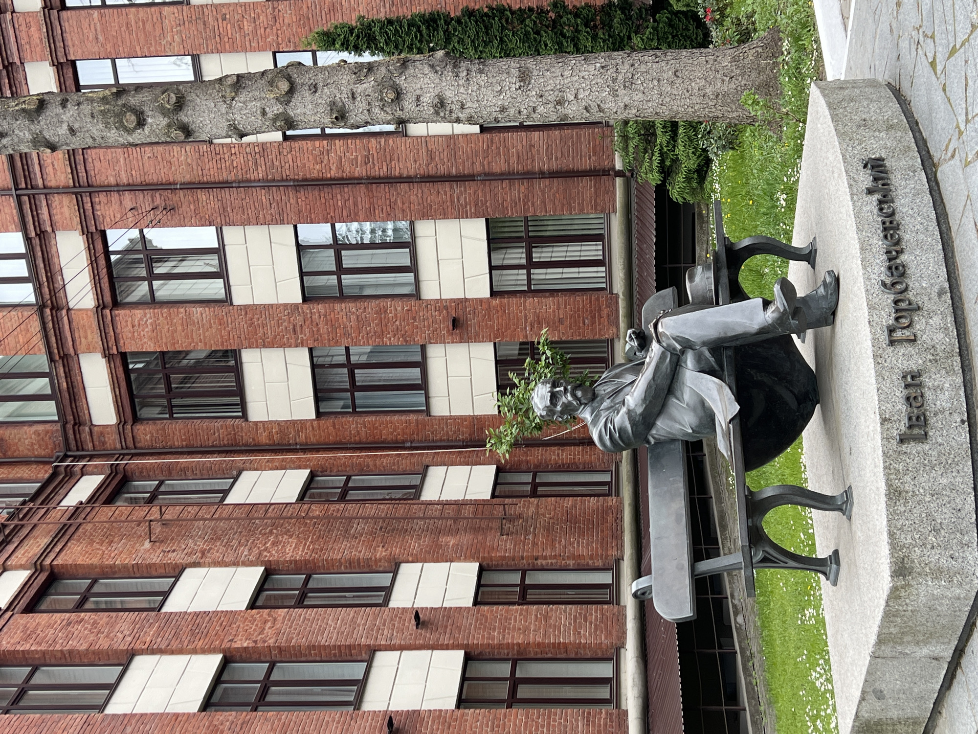 A photograph depicts a monument of Ivan Horbachevsky, a Ukrainian chemist and hygienist, located in Ternopil, Ukraine. The monument features a bronze statue of Horbachevsky seated on a bench. The statue is situated on a gray stone plinth with the name "Іван Горбачевський" inscribed on the side in Ukrainian. Behind the statue is a multi-story brick building with windows. There is a tree and greenery surrounding the monument. The photo is taken from a slightly low angle.