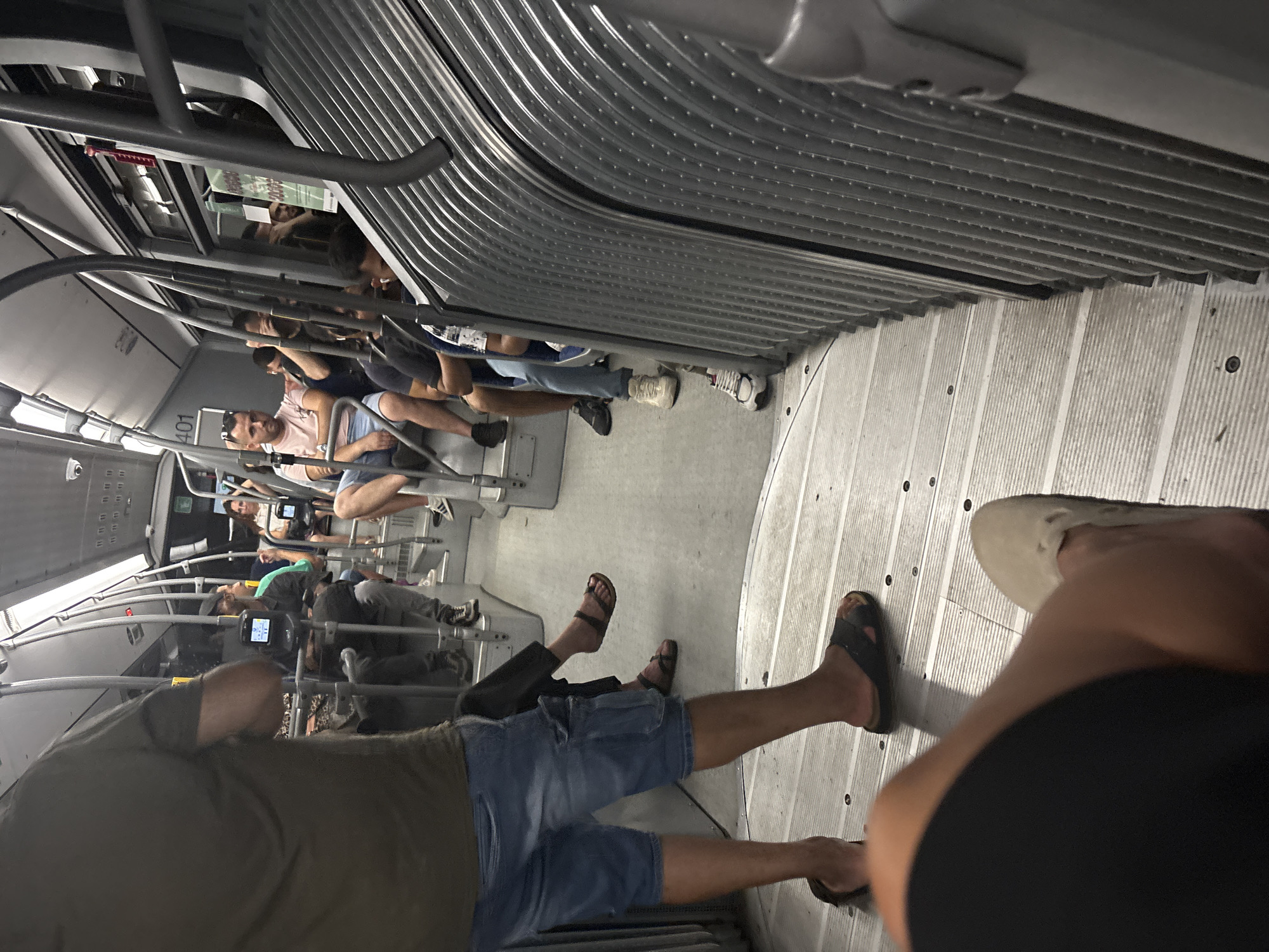 The image depicts the interior of a crowded public transportation vehicle, likely a bus or tram, with passengers seated and standing. The scene appears to be a typical urban commute. There are individuals of various ages and genders visible, dressed in casual clothing. The environment is utilitarian, with metal poles for support and ribbed seating or wall panels. The lighting suggests it is either daytime or a well-lit interior. The specific location of Venice, Italy, cannot be determined from the visual information provided in the image.