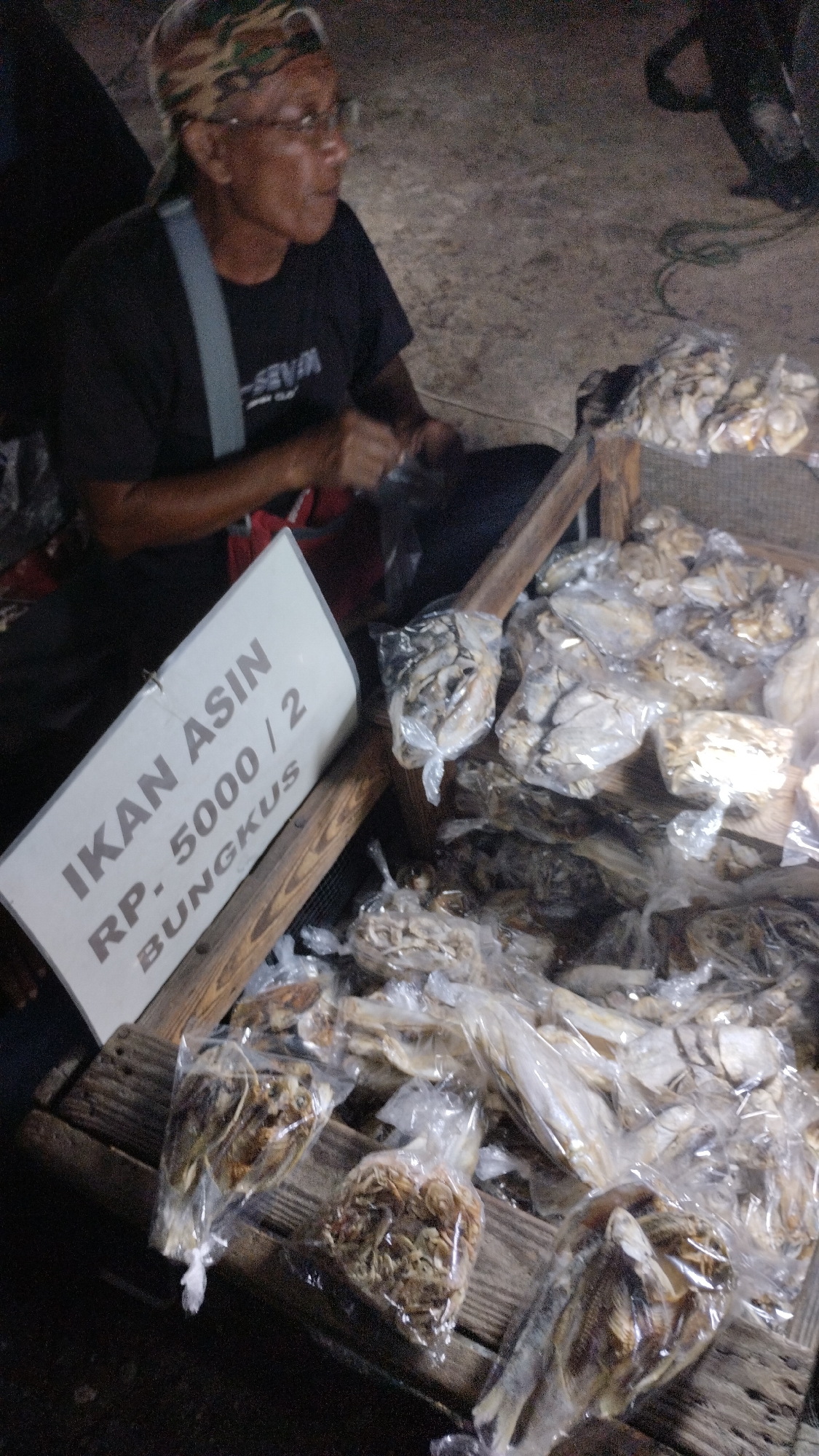 In Sambungrejo, Indonesia, a person wearing glasses and a camouflage bandana sits next to a display of dried fish. The person is wearing a black t-shirt with a gray strap across their chest. They are looking to the right, with a slight smile.
The dried fish are arranged in plastic bags on a wooden crate. Some bags contain whole dried fish, while others contain smaller, shredded pieces of fish. A white sign with text in Indonesian is propped up on the crate. The sign reads "IKAN ASIN RP. 5000 / 2 BUNGKUS," which translates to "Salted Fish Rp. 5000 / 2 Packs."
The scene appears to be outdoors, possibly at a market or roadside stall, given the natural lighting and the informal setup. The time of day is unclear, but the lighting suggests it could be evening or early morning. The overall mood is relaxed and transactional.