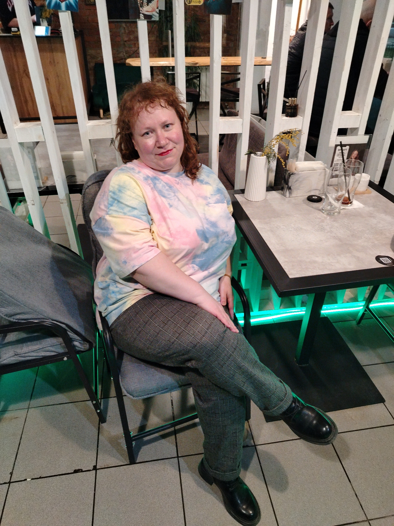 A medium shot captures a woman with reddish-brown curly hair, wearing a colorful tie-dye t-shirt (pink, yellow, blue) and grey plaid trousers, seated at a table in what appears to be a cafe. She is looking directly at the camera with a slight smile and red lipstick. Her left leg is crossed over her right, and she wears black, sturdy shoes.
The setting is indoors, featuring white slatted partition walls that partially obscure the background, revealing a brick wall and other patrons. The table in front of her has a light grey surface and a black frame. On the table are a white ribbed vase holding yellow floral sprigs, a white rectangular napkin dispenser, a small black diffuser bottle, two empty drinking glasses, a small white cup, and a black coaster. A menu or informational card on the table displays "BR CAFE" and a QR code. Green LED strip lights illuminate the area under the table and along the base of the partition walls, suggesting an evening or night-time setting. The floor is tiled with light grey squares. Another chair with a grey fabric cushion is partially visible to her left. In the background, a blurred figure is seated behind the partitions, and a sign with "PUSH O" is faintly visible. The woman appears relaxed, posing for the photograph in a casual, contemporary cafe environment in Chaikovsky, Russia.