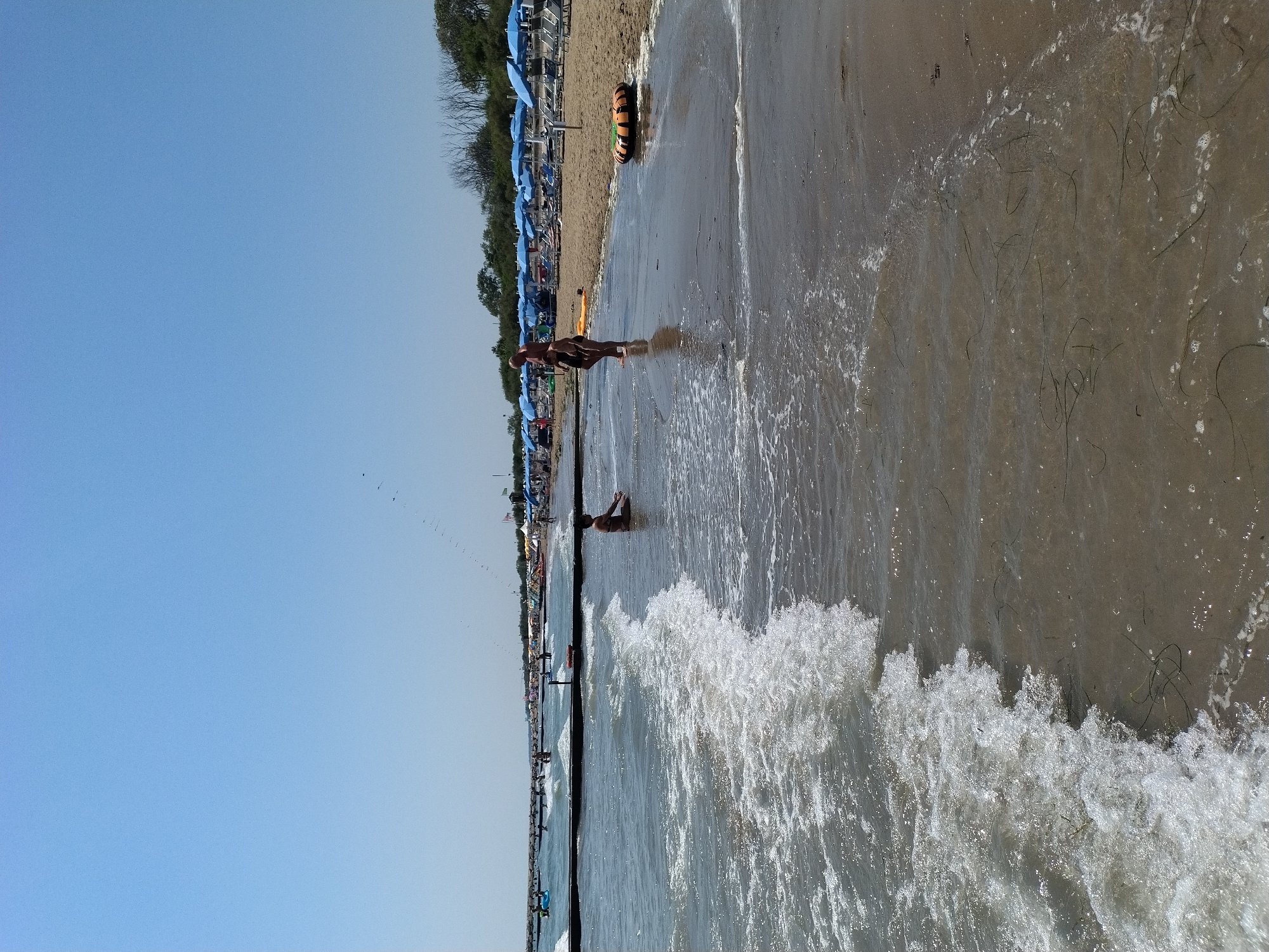 This image depicts a section of the beach in Caorle, Italy, on a clear, sunny day. The foreground is dominated by gentle waves washing onto the sandy shore. In the middle ground, a young person is seated in the shallow water, and further back, an adult and a child stand on the wet sand. The background reveals a bustling beach scene with numerous blue umbrellas providing shade over lounge chairs, with trees and vegetation lining the edge of the beach. The sky is a vast expanse of clear blue, with a faint, linear formation of birds visible high above.