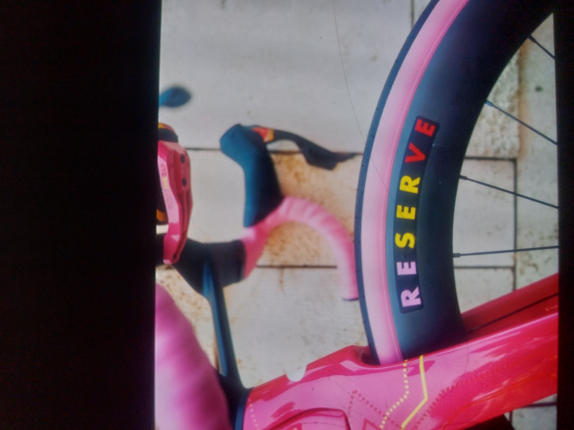 The image shows a close-up of a pink bicycle with "RESERVE" written vertically on the tire in colorful letters. The bicycle's handlebars are wrapped in pink tape and have dark-colored brake levers. The bicycle is resting on a light-colored surface that appears to be concrete or stone. The location and city of Tsarevo, Bulgaria are not discernible from this image.