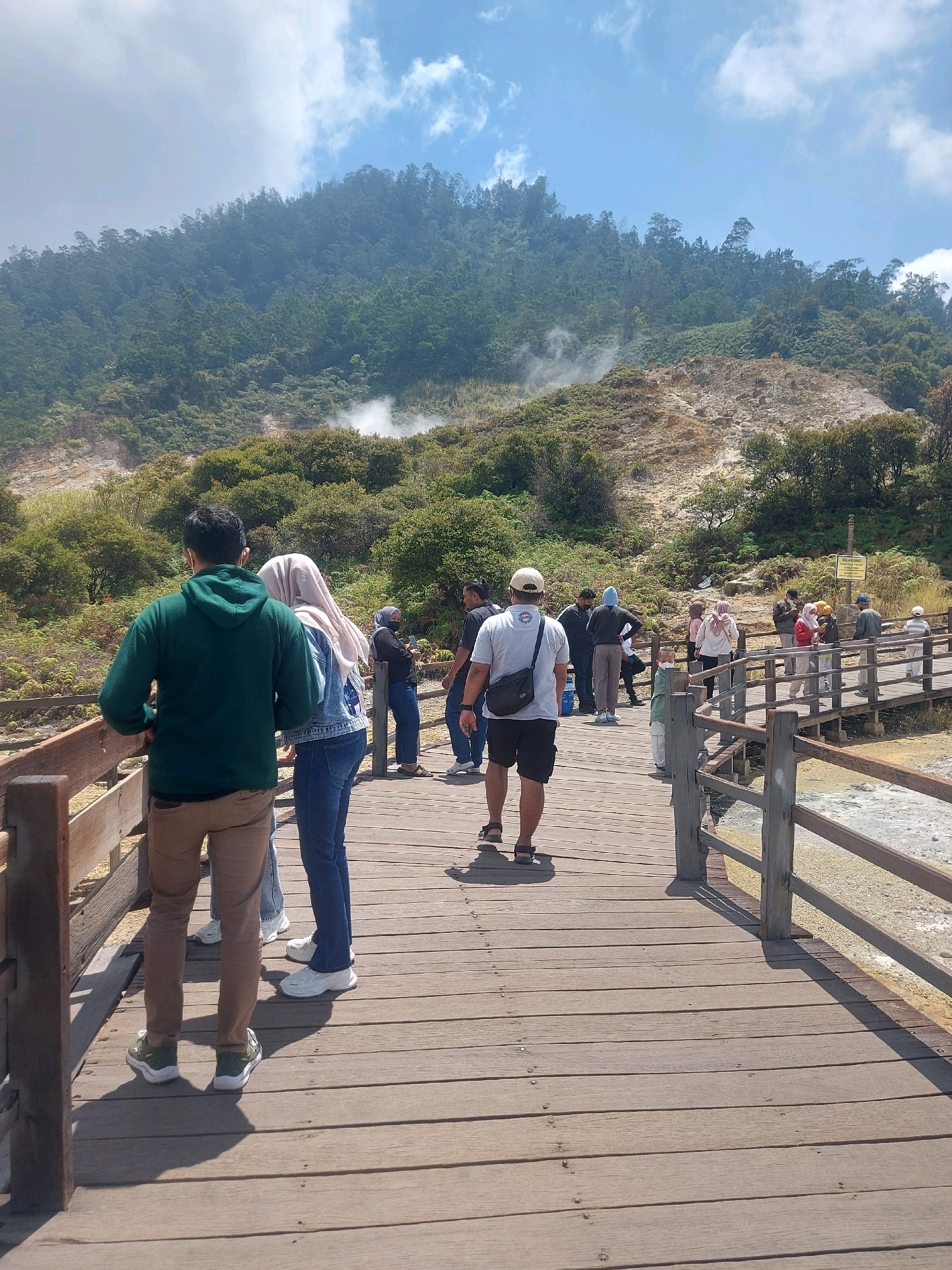 The image depicts a scene at the Sikunang Crater in Dieng, Indonesia, a geothermal tourist attraction. A wooden boardwalk, constructed from planks of wood, serves as a pathway for visitors. The boardwalk winds through a landscape characterized by volcanic activity. In the background, a forested hillside rises, with plumes of white steam emanating from the crater, indicative of its geothermal nature. The ground surrounding the boardwalk appears barren in places, with some patches of vegetation.
Several individuals are present on the boardwalk, dressed in casual attire. On the left, a person in a dark green hooded sweatshirt and tan pants stands, facing away from the camera, looking towards the crater. Next to them, a person wearing a light-colored hijab and a denim jacket, also facing away, stands with a backpack. Further down the boardwalk, more visitors are seen walking and observing the scenery. The weather appears to be sunny with some clouds in the sky. The overall atmosphere suggests a tourist excursion to a natural wonder.