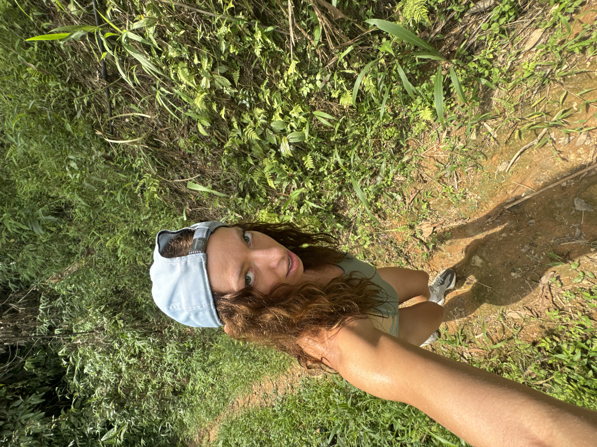 A woman stands on a dirt path, looking up at the camera. She wears a light blue baseball cap, a green top, and gray and white running shoes. Her arm extends towards the camera. The path is lined with lush green vegetation. The location is Suối Giàng, Vietnam.