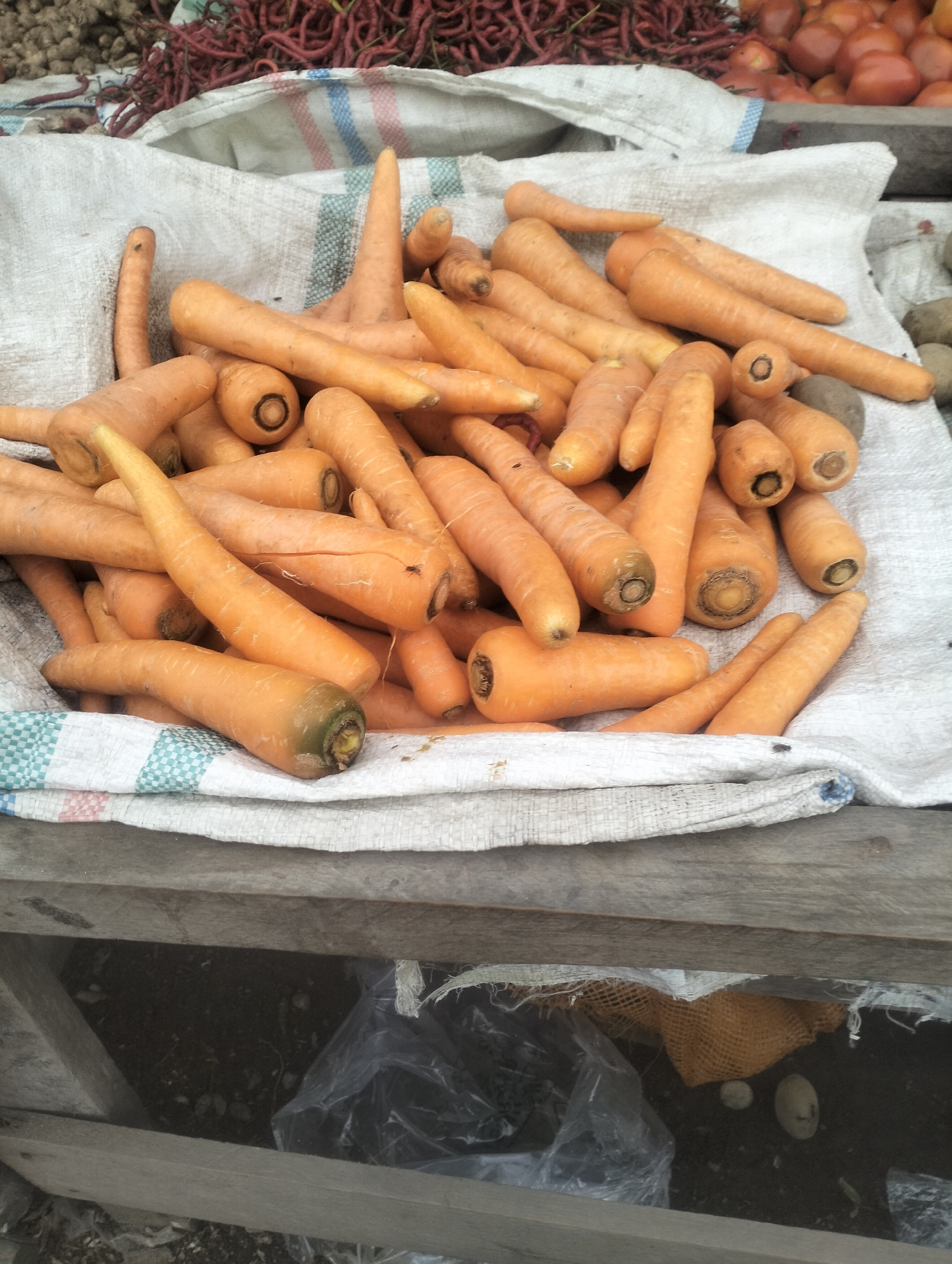 The image shows a pile of carrots on a white, woven sack, likely at an outdoor market in Dumai City, Indonesia. The carrots are predominantly orange, with some showing green tips. They vary in size and shape, with some appearing smooth and others slightly irregular. The sack has horizontal stripes of red, blue, and white, and is resting on a wooden surface. In the background, out of focus, are other vegetables, including what appear to be tomatoes and possibly potatoes, as well as a bundle of red chili peppers. The scene suggests a vendor’s stall or a display of fresh produce.