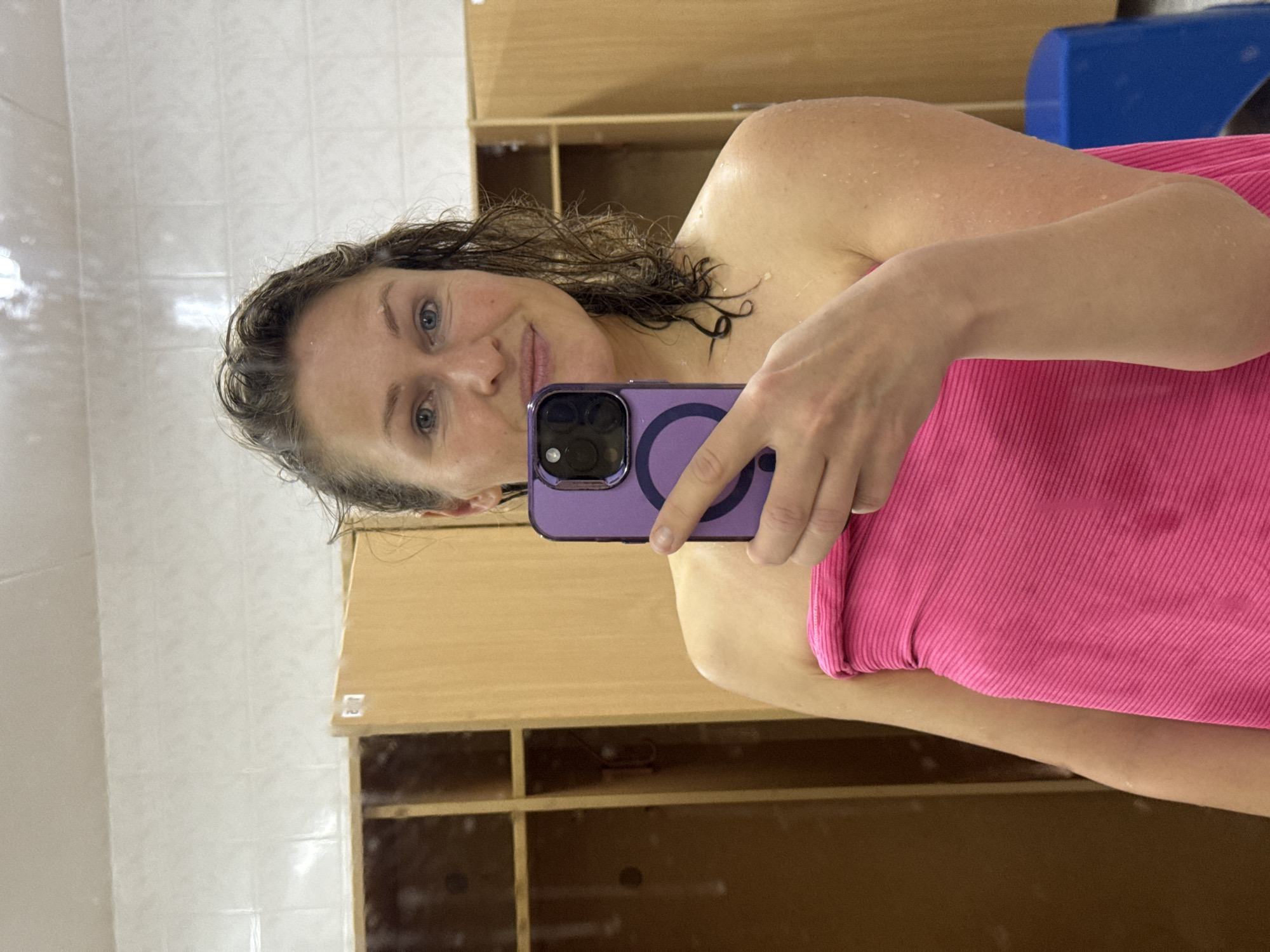 A woman takes a selfie in what appears to be a changing room or spa area in Chaikovsky, Russia. She is positioned vertically in the frame, with her head and upper torso visible. Her dark, wet hair is loosely styled, and her face shows a calm expression with a slight smile, revealing blue eyes. Water droplets are visible on her skin, suggesting she has recently been in water, possibly after swimming or a shower. She is draped in a bright pink ribbed towel or wrap around her upper body.
In her right hand, she holds a purple smartphone with a prominent circular design on its back, likely indicating a MagSafe feature. The phone is pointed towards her, indicating she is using it to capture the image.
The background consists of light-colored tiled walls on the left and upper parts, and wooden lockers or shelving units on the right and lower sections. A blue object, possibly a container or piece of equipment, is partially visible in the upper right background. The overall impression is that of an indoor, functional space, commonly found in gyms, pools, or spas. No visible text is discernible in the image.