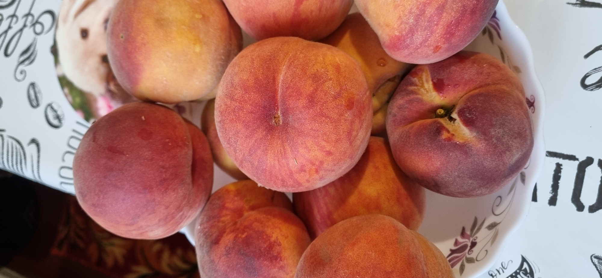A still life of peaches in a white bowl is depicted. The bowl is patterned with flowers and a floral border. The peaches are a mix of red and yellow hues, with some showing more red blush than others. A soft, diffused light illuminates the scene, casting subtle shadows. The background is out of focus, but appears to be a patterned tablecloth and possibly a hint of a light-colored surface. The overall impression is one of abundance and freshness. The location in Blizhnee, Ukraine, is not evident from the image.