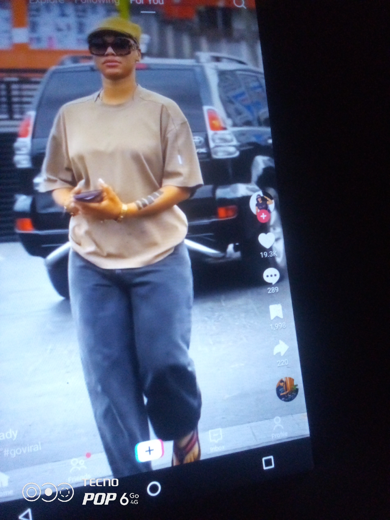 The image shows a woman standing in front of a dark SUV. She is wearing a light brown t-shirt, dark blue jeans, and a green flat cap. She is also wearing sunglasses and has jewelry on her wrists. The woman is holding a dark object in her hands, which appears to be a phone. The background is blurred, but it seems to be an outdoor setting, possibly a street. There are social media icons and text overlaid on the image, suggesting it is a screenshot from a social media platform. The text "#goviral" is visible, indicating a desire for widespread attention. The presence of a Tecno Pop 6 4G logo suggests the image was captured on a Tecno Pop 6 4G phone. The scene is not specifically identifiable as being in Howayi, Nigeria, or any other particular city.