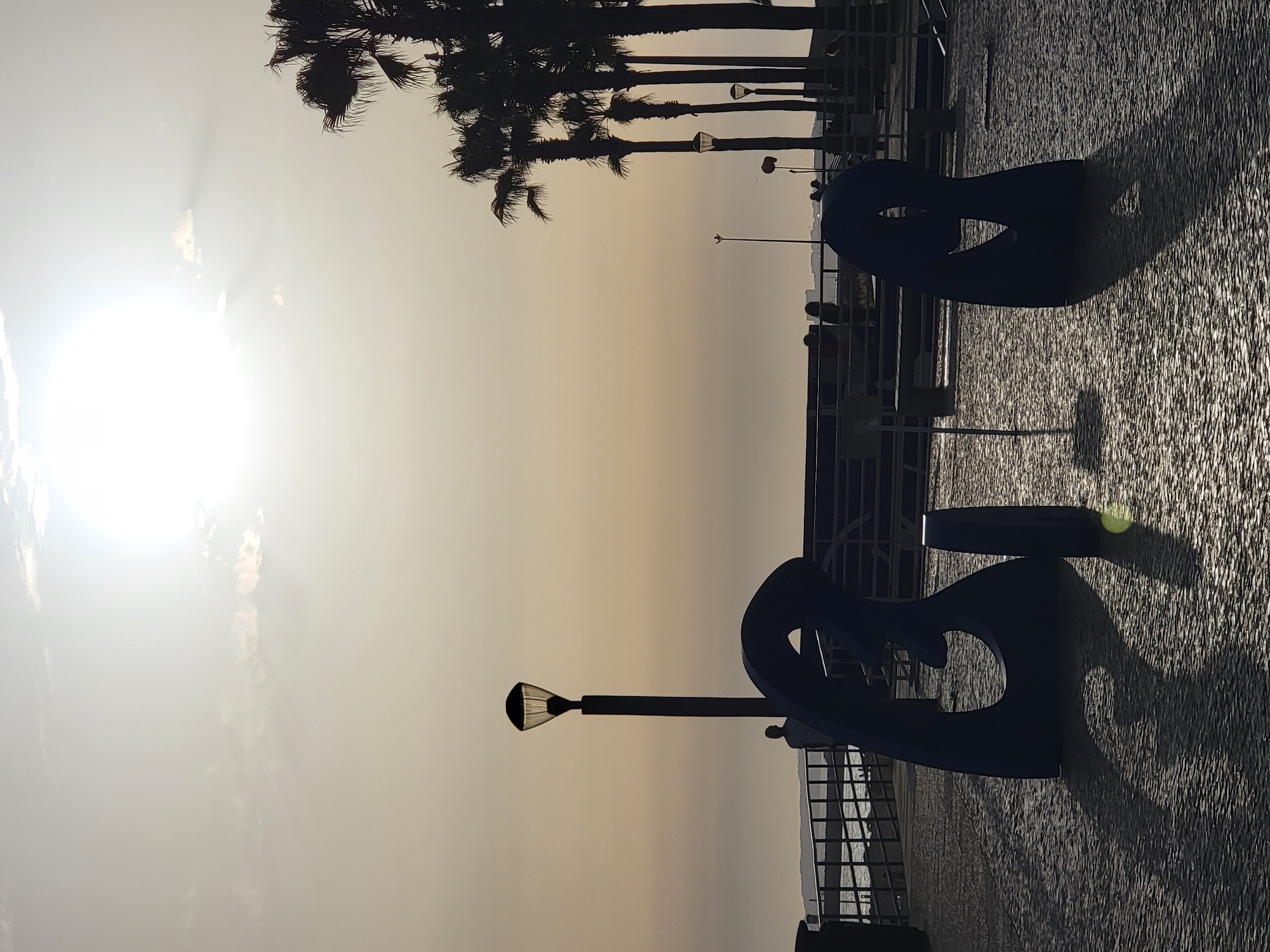 The image depicts a scene in Quarteira, Portugal, at dusk. The sky is a muted yellow-orange, with the sun prominently visible as a bright, diffused orb near the top of the frame, casting rays through a few wispy clouds. Silhouetted against the sky are several tall palm trees on the right side of the image. In the foreground, two abstract, dark blue sculptures are positioned on a patterned paved surface. The sculpture on the left is larger and more complex, while the one on the right is smaller and simpler. Long, distorted shadows of these sculptures are cast across the pavement due to the low angle of the sun.
In the middle ground, a lamppost with a distinctive geometric shade stands to the left of the frame. Beyond it, a railing runs along a walkway, with the ocean visible in the distance. Several figures are visible as silhouettes on the walkway, including one person standing near the lamppost and others further down, some seated on a bench. The environment appears to be a public promenade or plaza overlooking the sea. The lighting suggests the golden hour, creating a serene and slightly melancholic atmosphere.